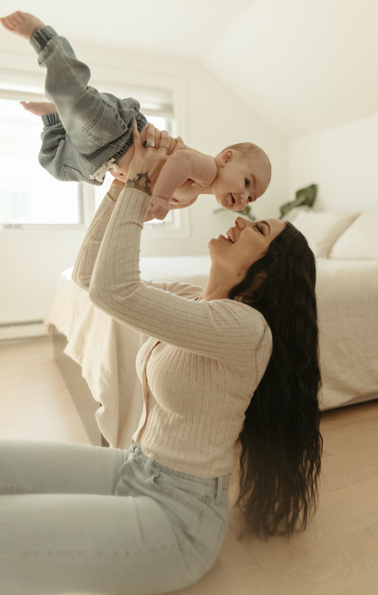 Woman lifting baby in a bright bedroom, smiling at each other, with a bed and window in the background.