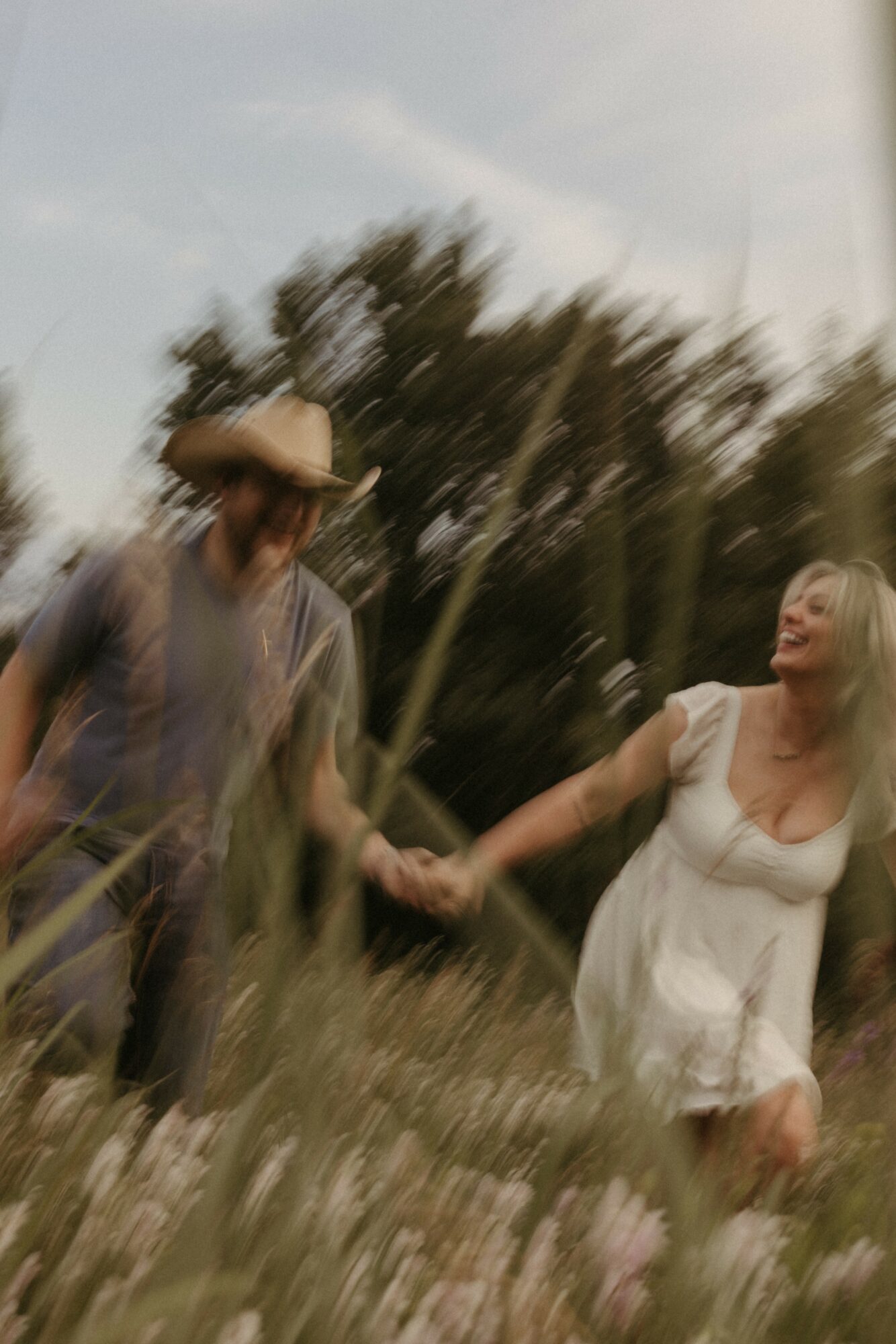 Two people holding hands outdoors, one wearing a hat and the other in a white dress, with trees in the background.