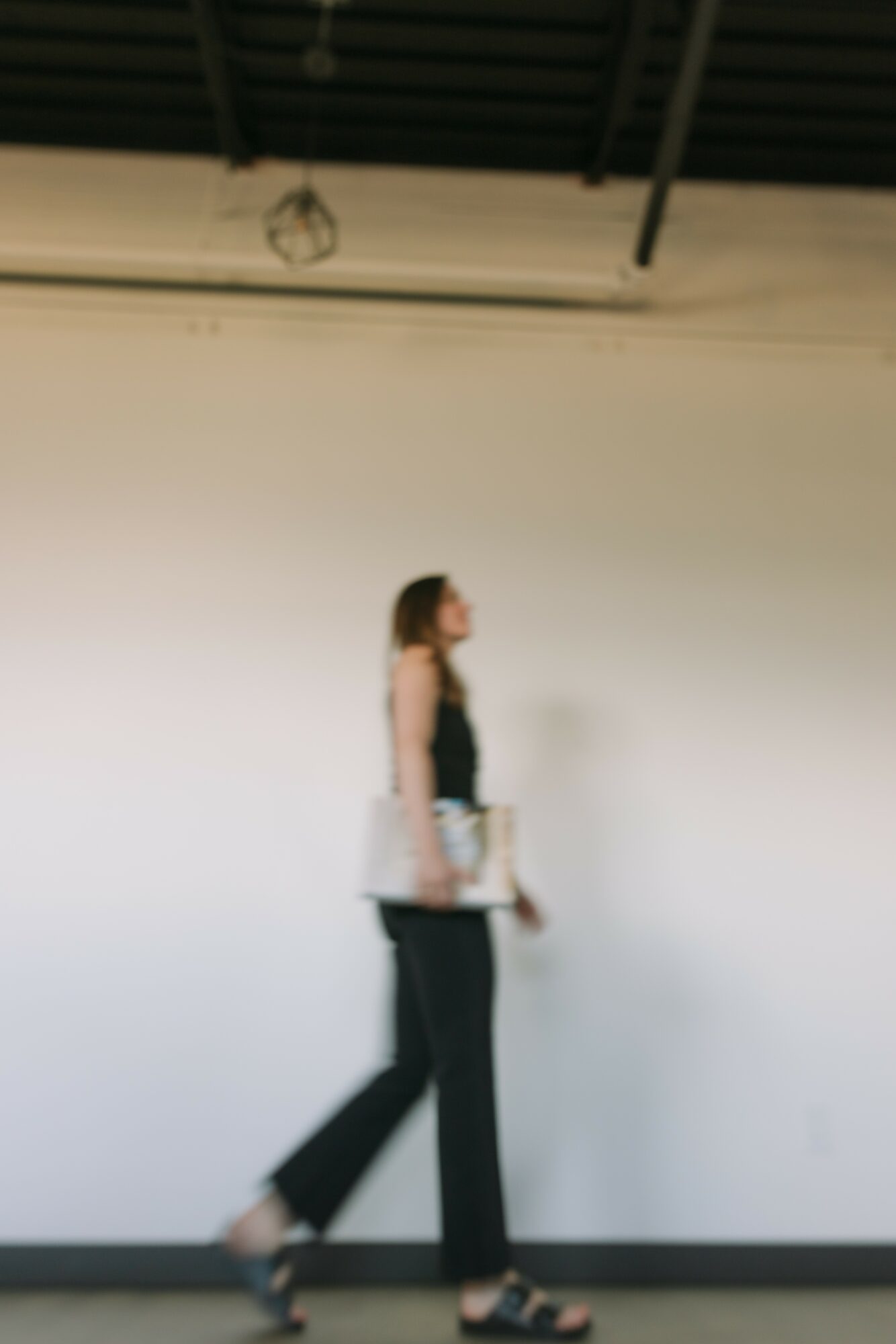 A woman walking with a bag in an indoor space with a high ceiling and white wall.