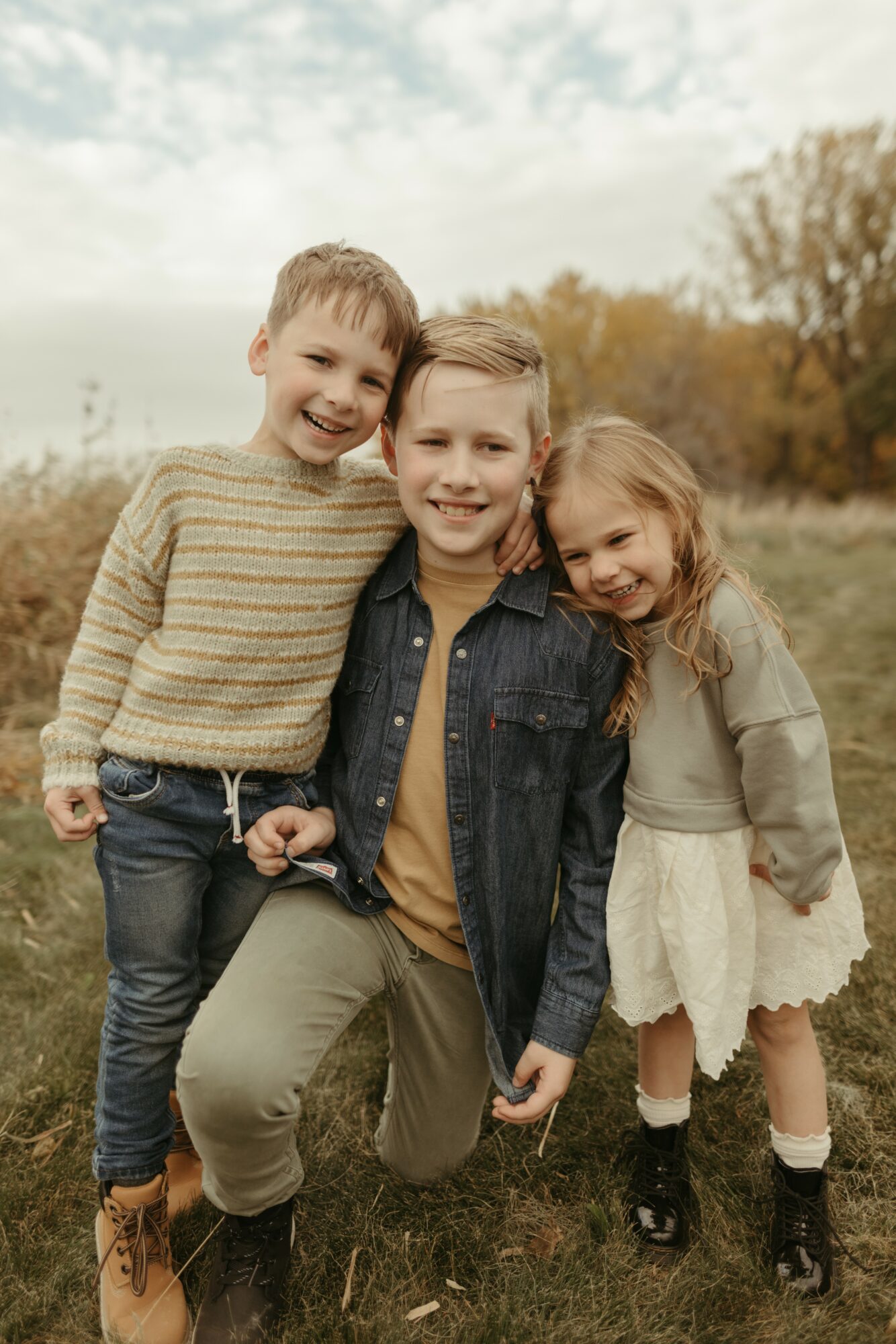 Three children outdoors, smiling, standing on grass with trees and sky in background.
