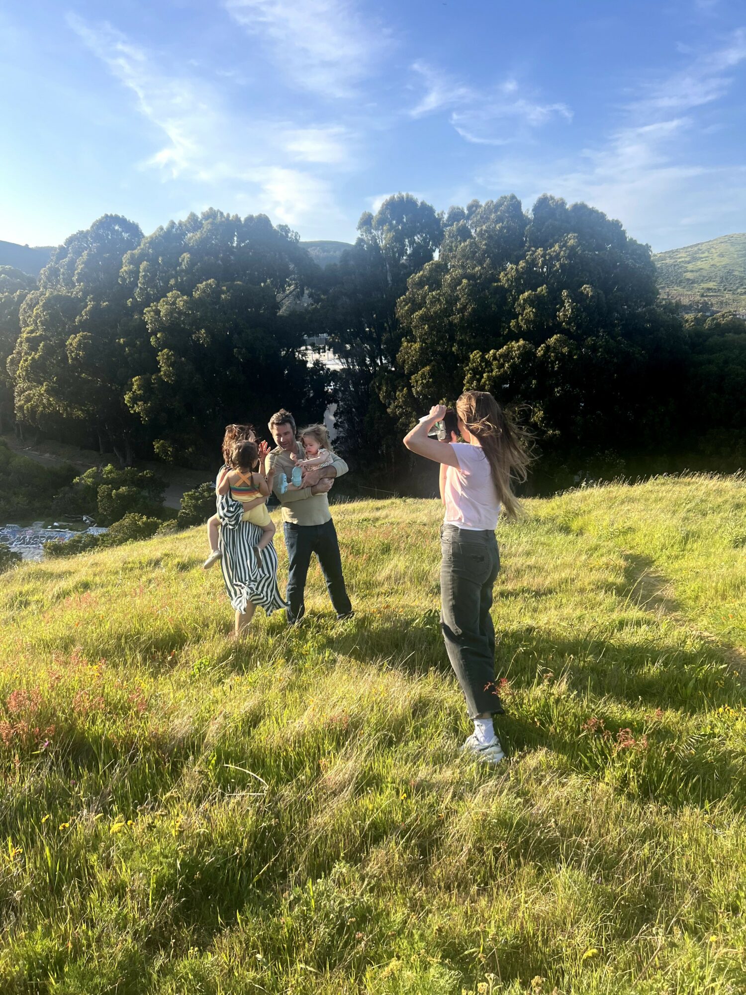 Four people standing on a grassy field with trees and blue sky in the background, two taking photos and two posing.