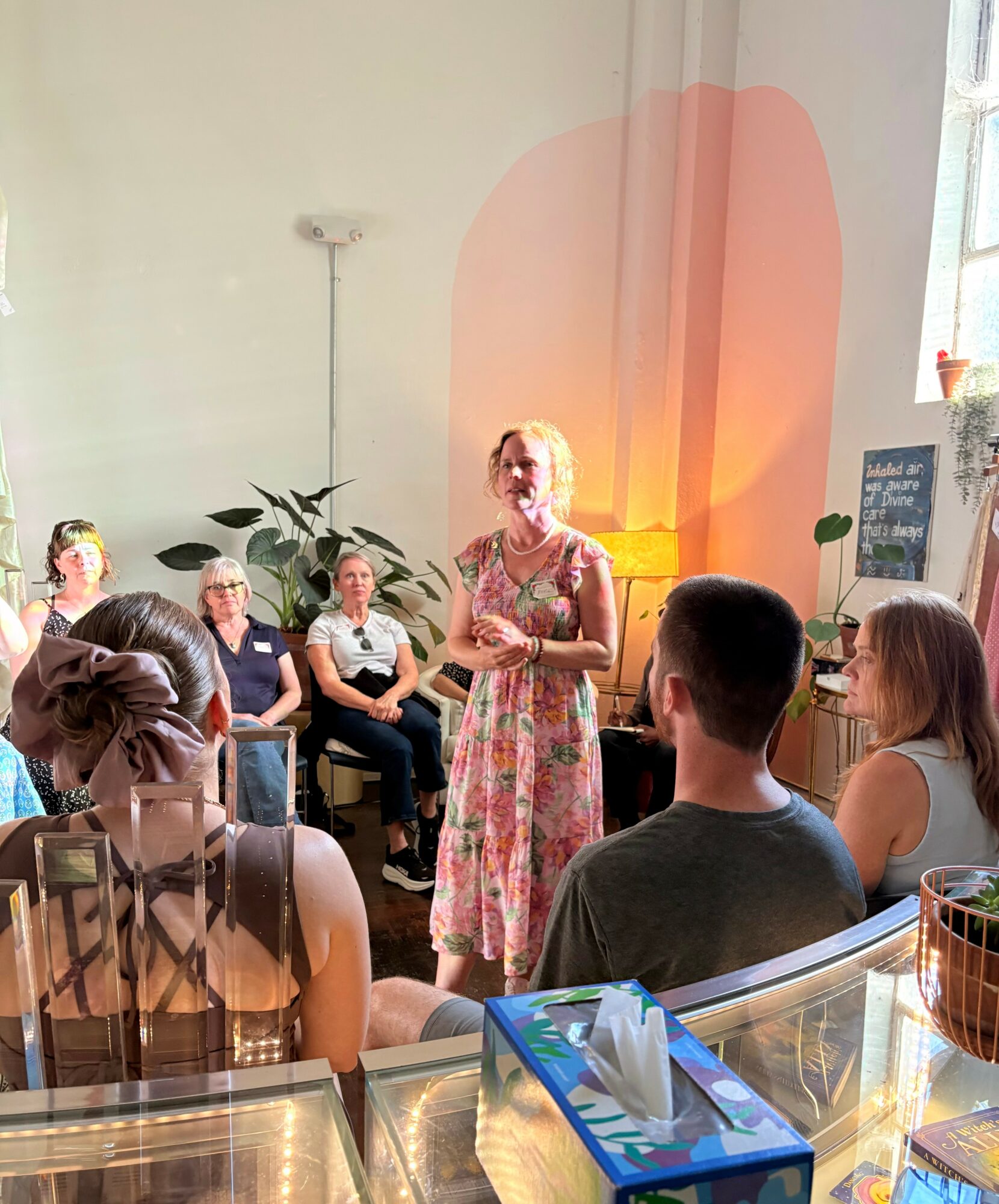 Woman speaking to a group in a bright room with sunlight, plants, and a chalkboard, seated on chairs and facing her.
