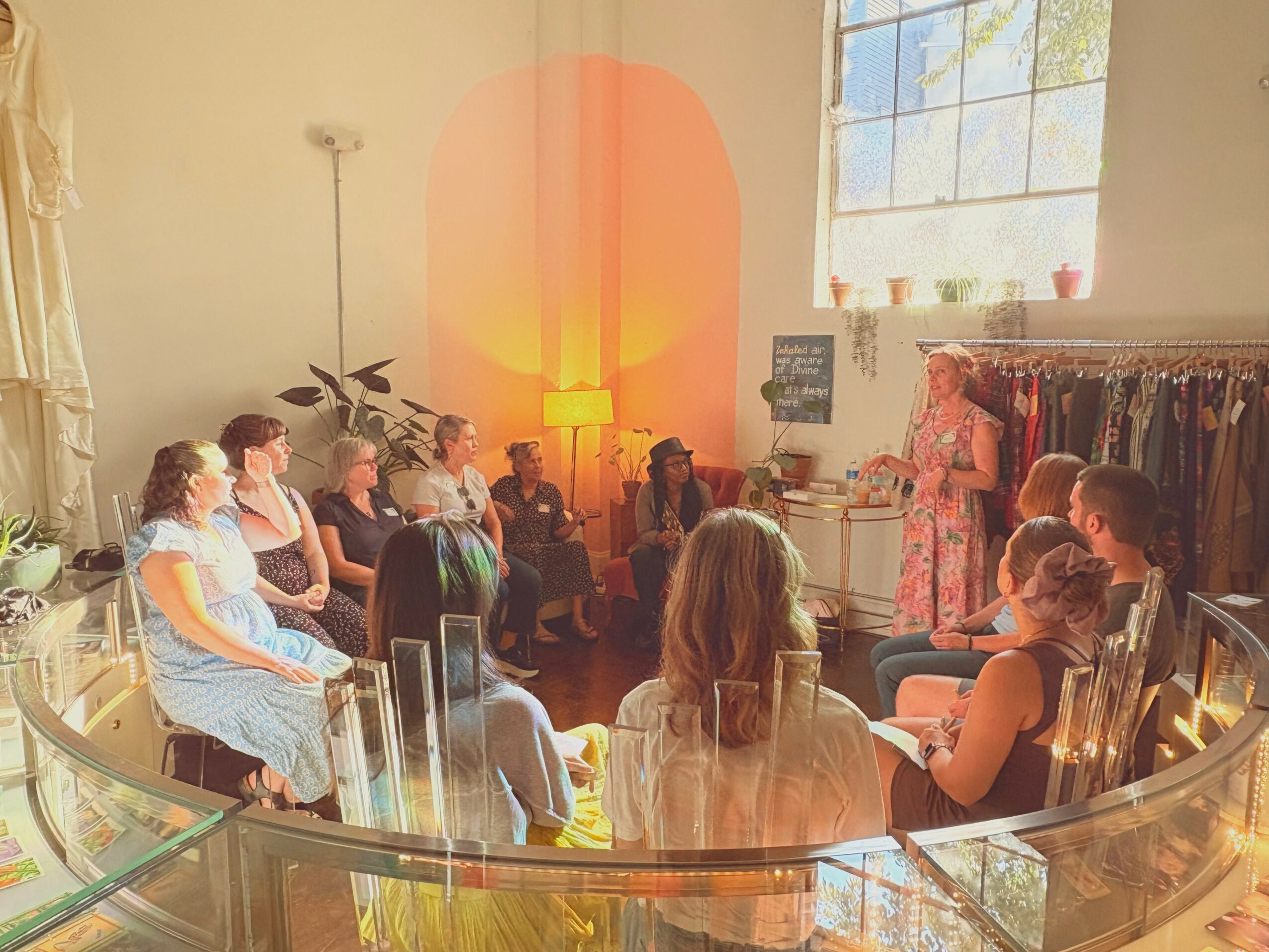 Group of people sitting in a circle indoors, with a window and clothing rack in the background.
