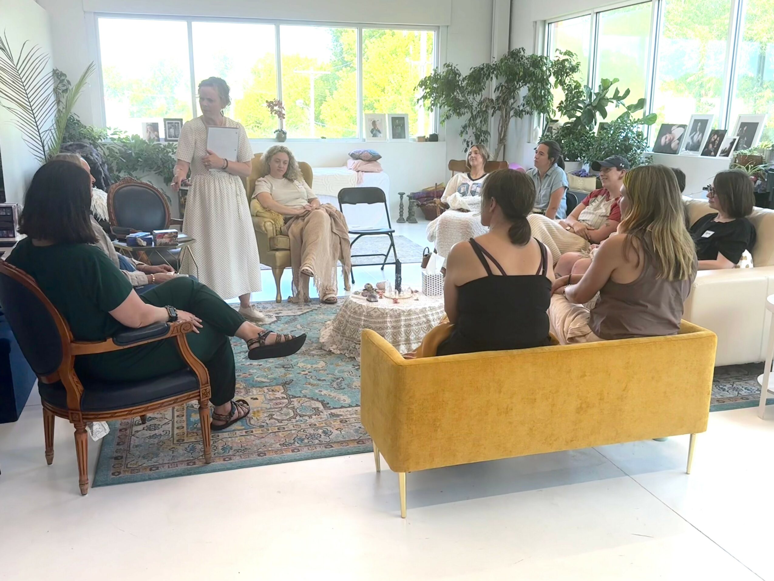 Group of women sitting and listening to a woman standing and speaking in a bright room with large windows and plants.