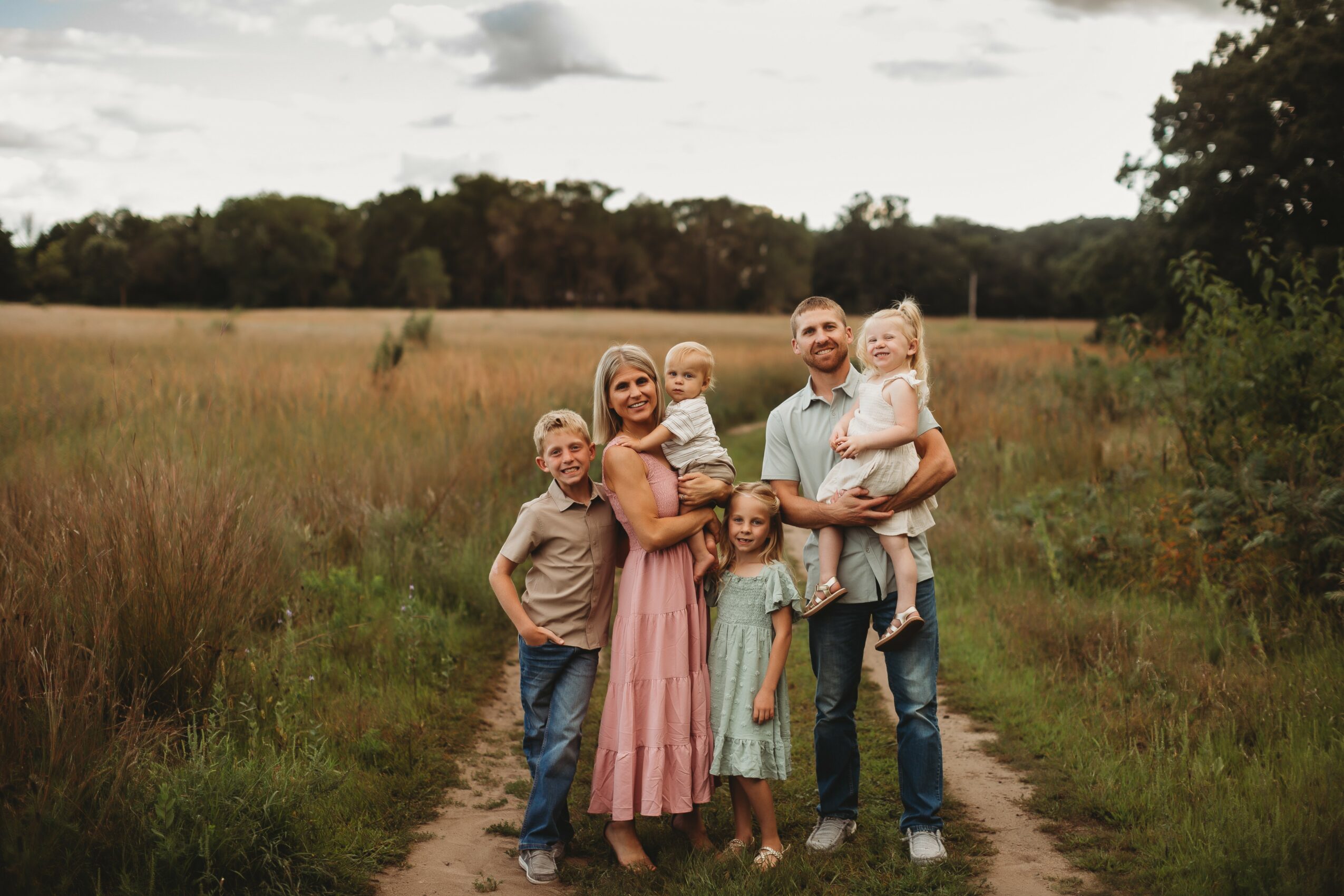 Family of six standing on a dirt path in a grassy field with trees in the background, smiling and posing for a photo.