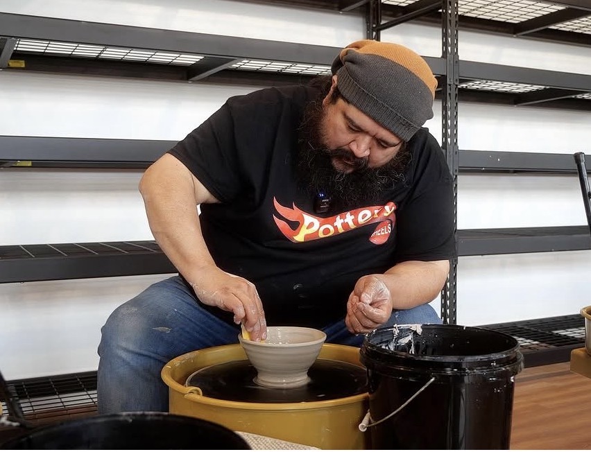 Person with a beard and gray beanie shaping pottery on a wheel, smiling, in a workshop setting.