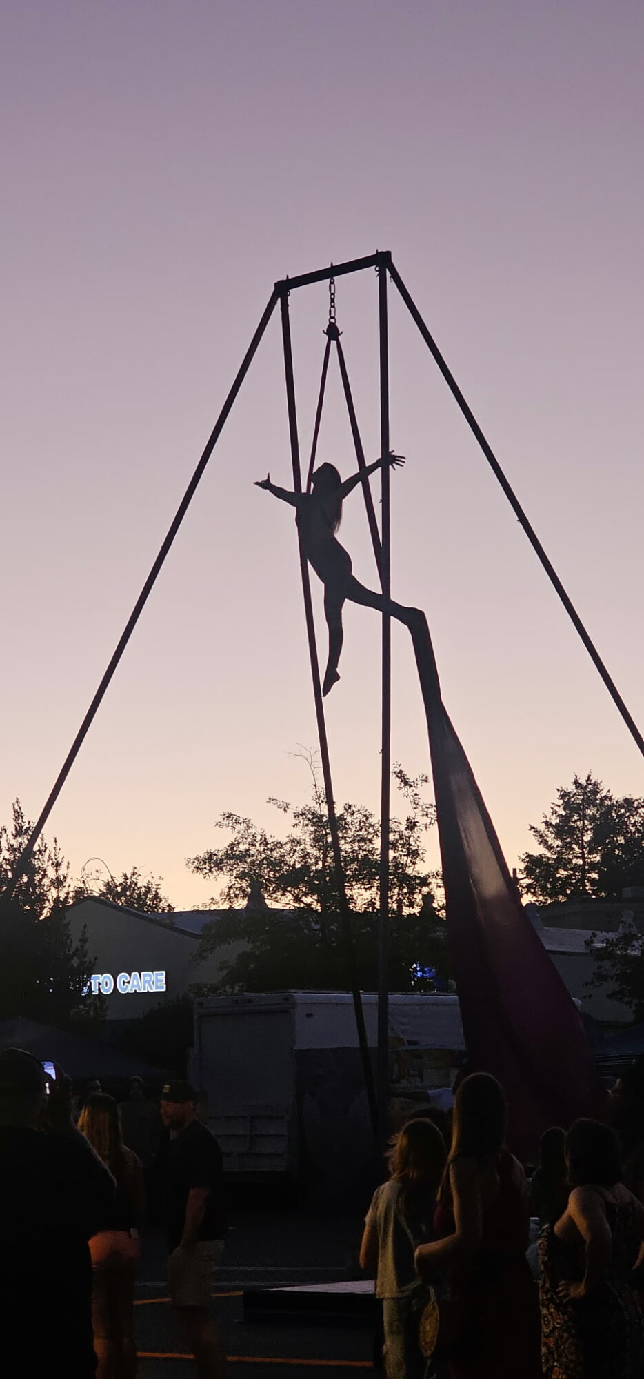 Person climbing a tall swing set structure against a sunset sky, with trees and people in the background.