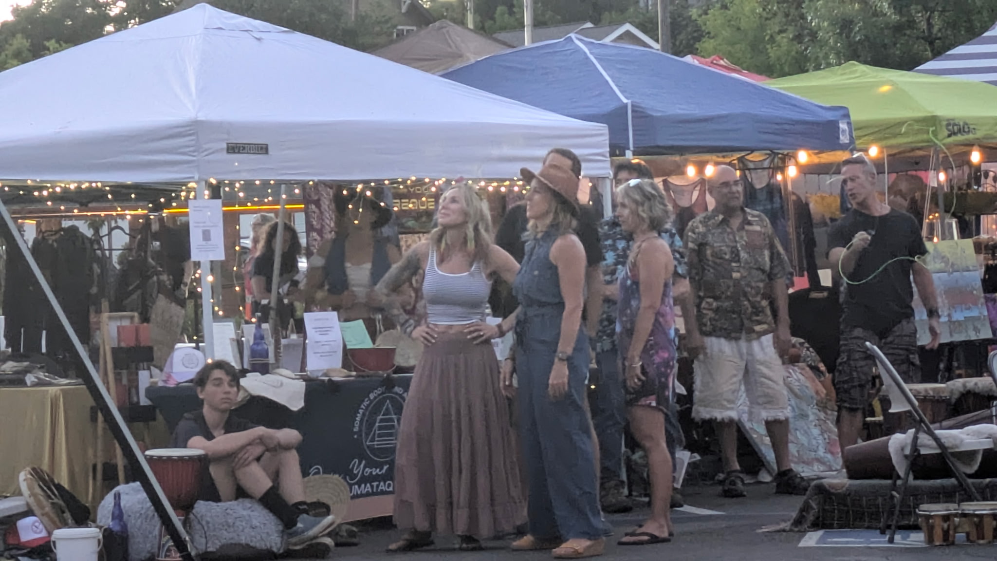People browsing market stalls under tents at an outdoor event, some sitting and some standing, in the evening.