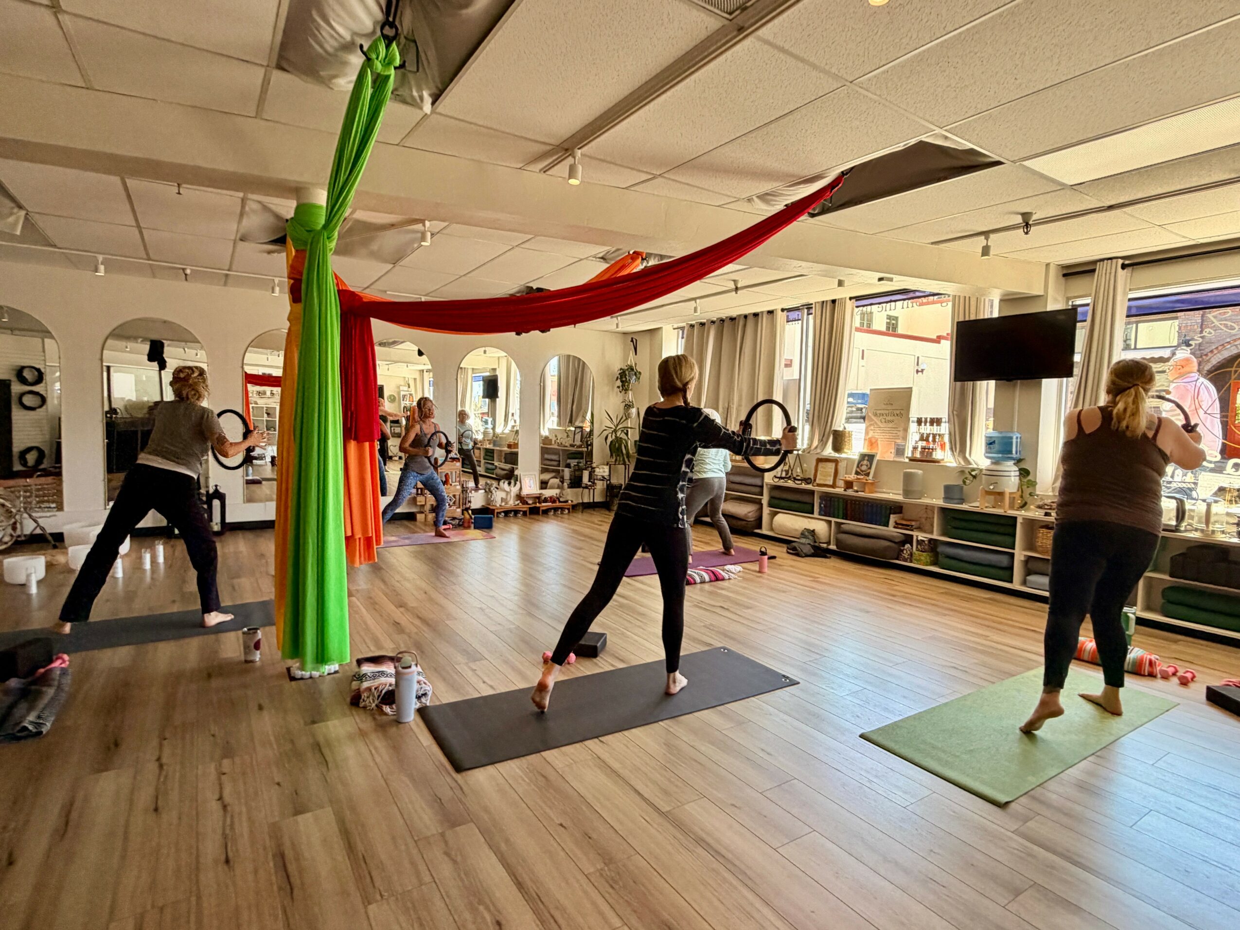 People participating in a fitness class in a bright room with large windows and wooden floors.