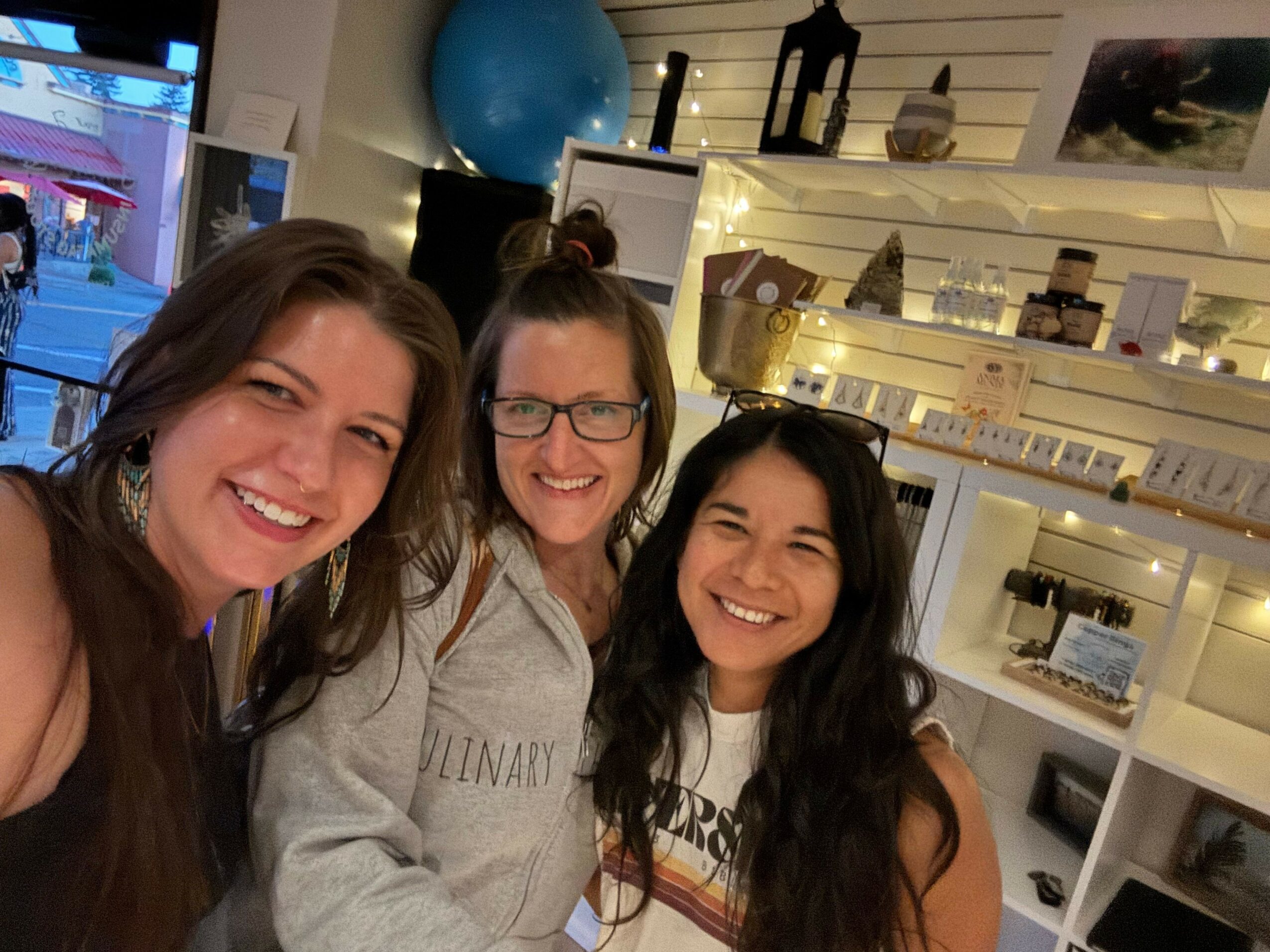 Three smiling women with long hair pose indoors, with shelves and a blue ball in the background.