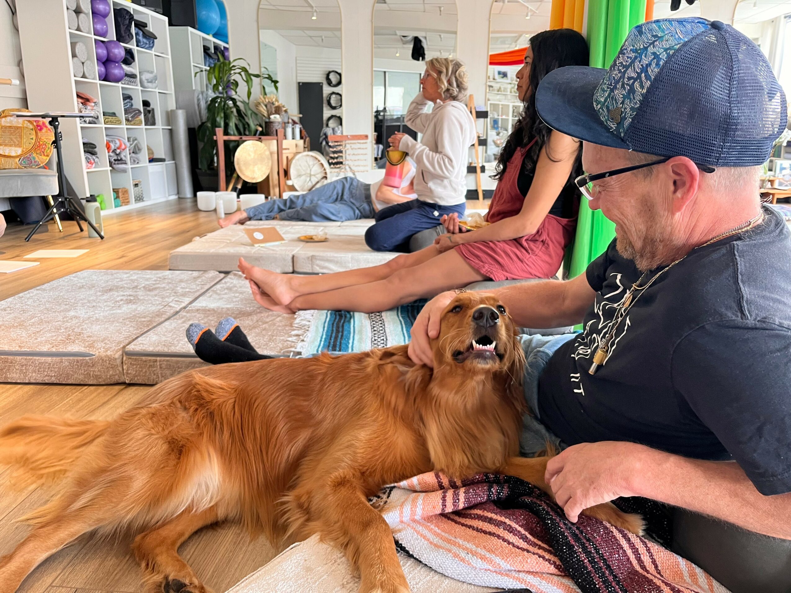 People sitting on the floor with a dog lying on its back, in a room with shelves and colorful objects.