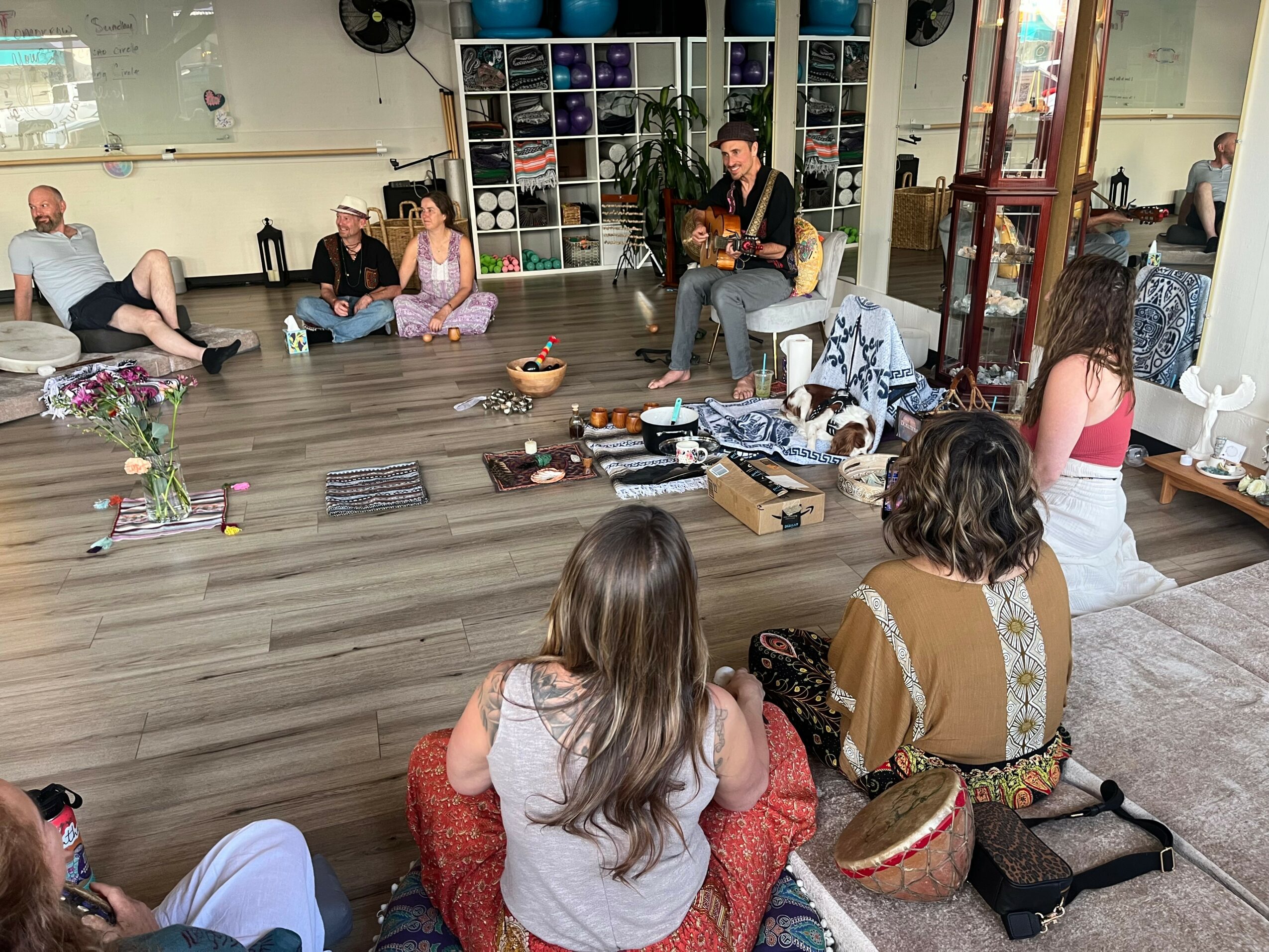 Group of people sitting on the floor and chairs in a room, some listening to a person playing guitar, with shelves and plants in the background.