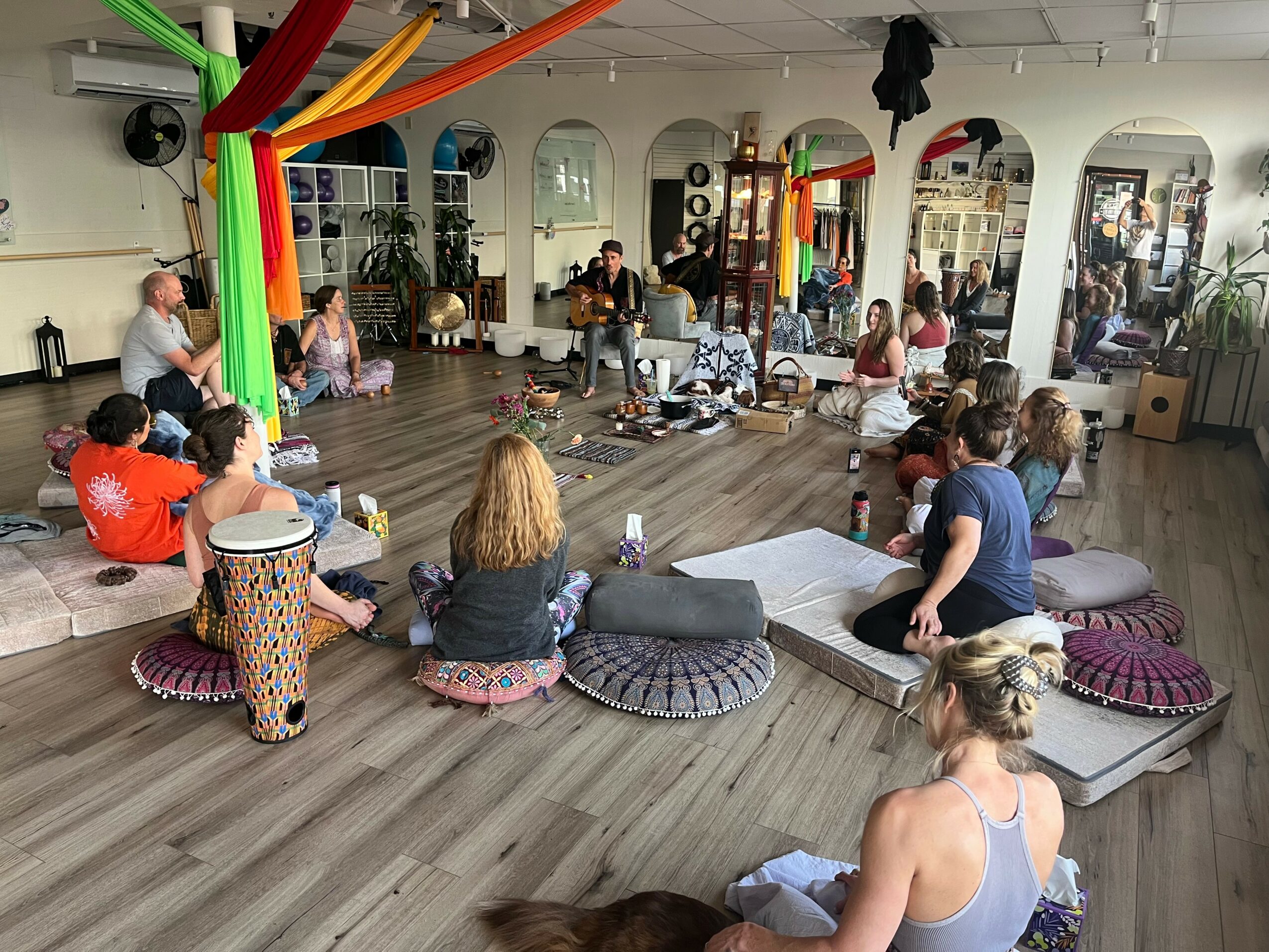 People sitting on cushions and mats in a spacious room with colorful decorations and large windows.