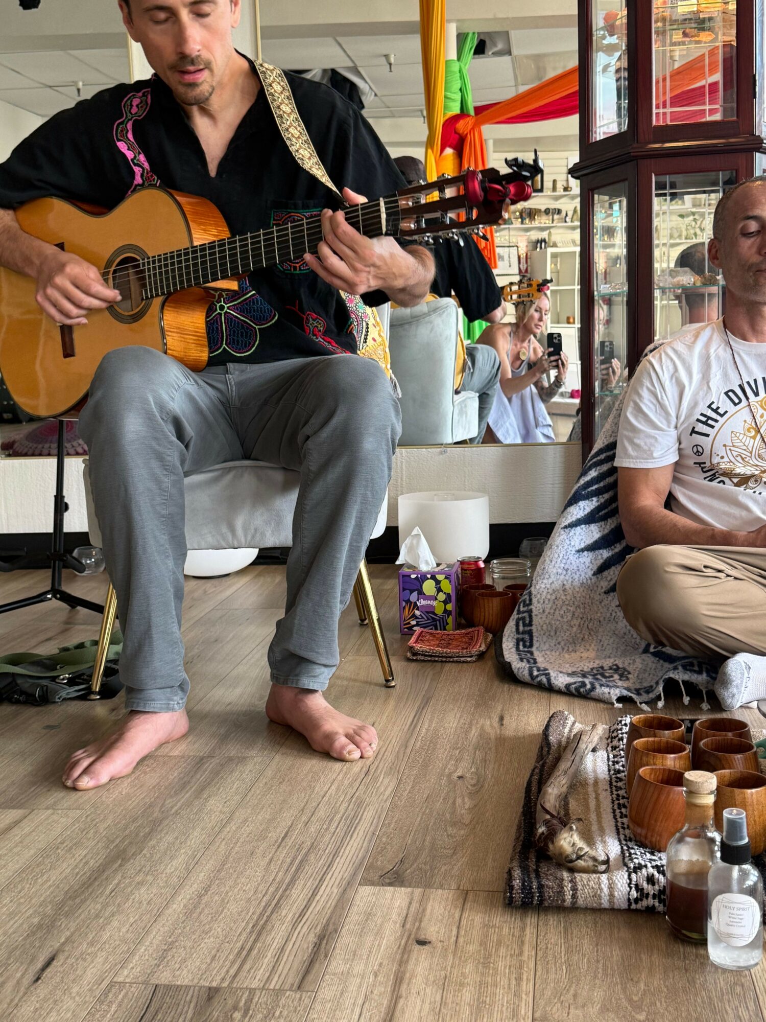 Person sitting on the floor playing guitar, with bottles and objects nearby, and people in the background in a room.