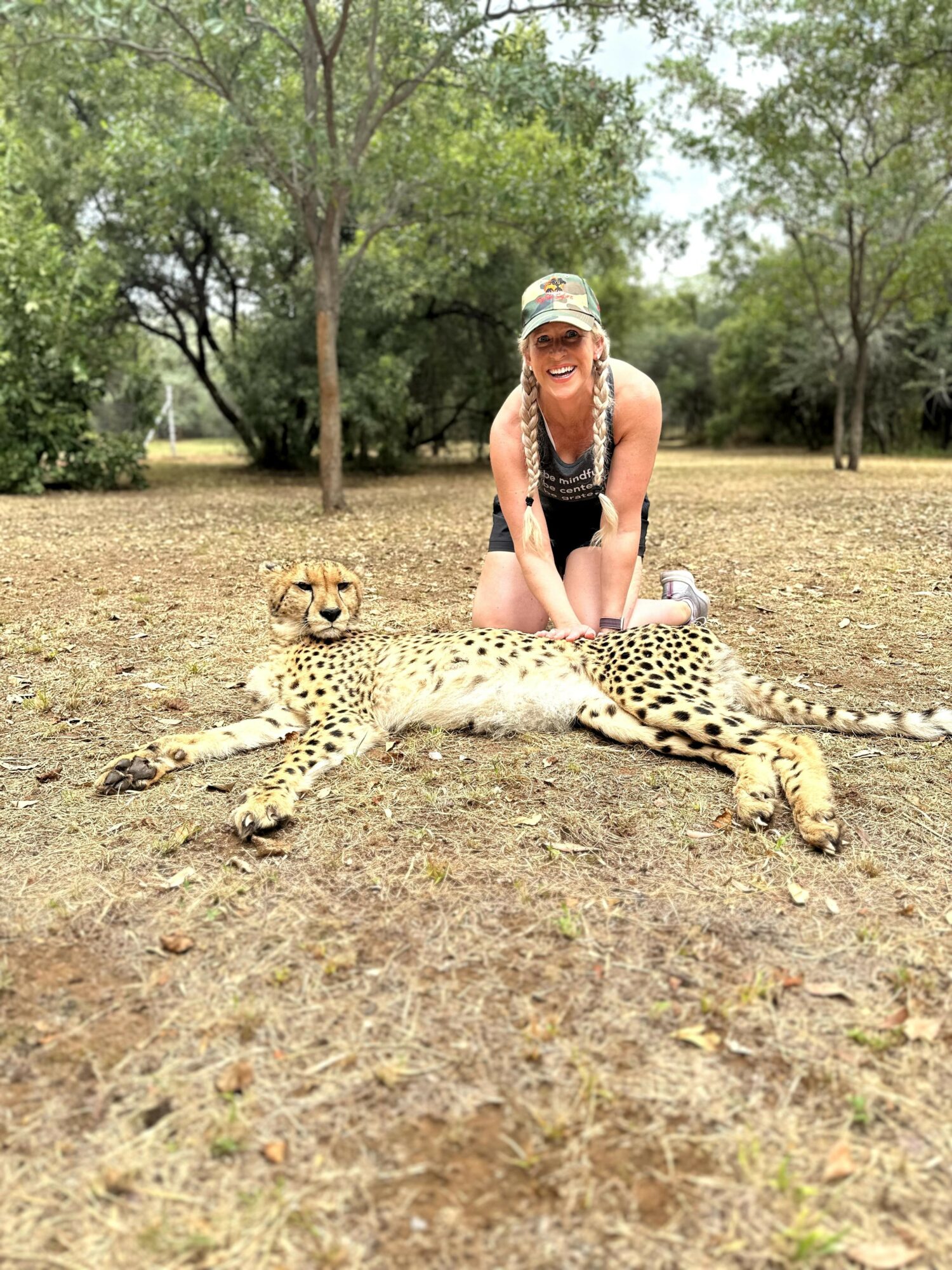 Person kneeling behind a lying cheetah in a park with trees, smiling and wearing a hat and sunglasses.