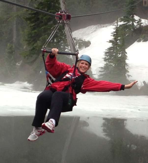 Person smiling on a zip line over water, wearing a helmet and red jacket, with trees and a waterfall in background.