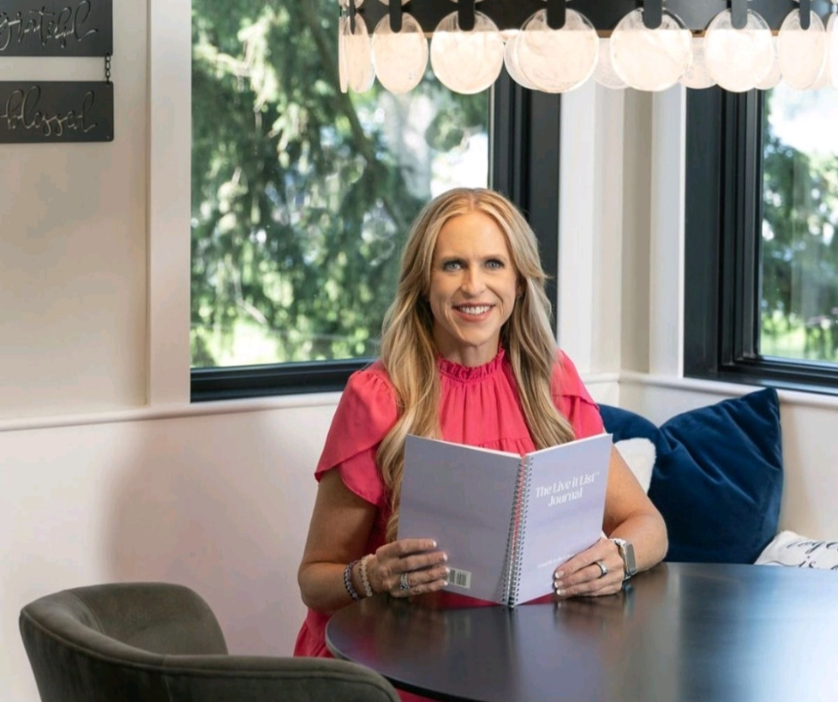Woman with long blonde hair in a pink blouse sitting at a table, holding a book, smiling, with windows and greenery outside