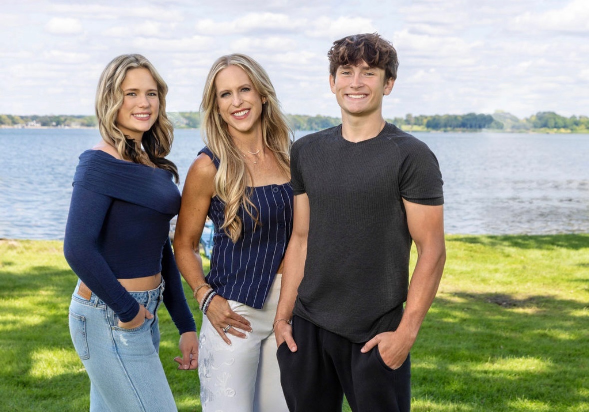 Three people standing outdoors near a body of water, smiling, with grass and trees in the background.