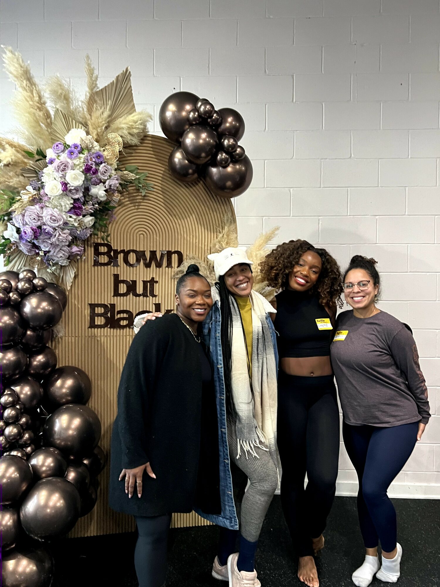 Four women standing in front of a decorated backdrop with balloons and flowers, smiling at the camera.