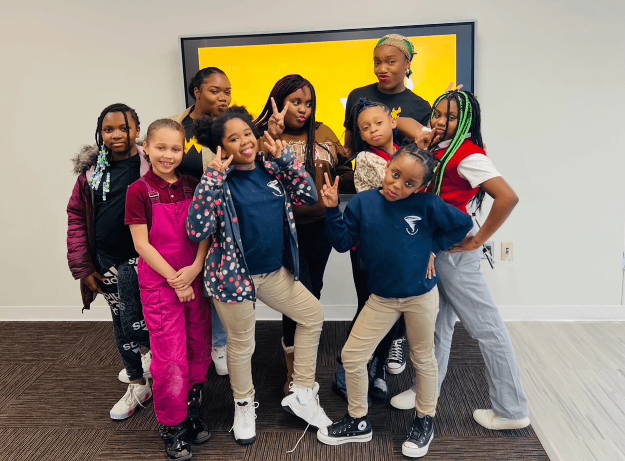 Group of children and adults posing together indoors in front of a yellow and black screen, smiling and making gestures.