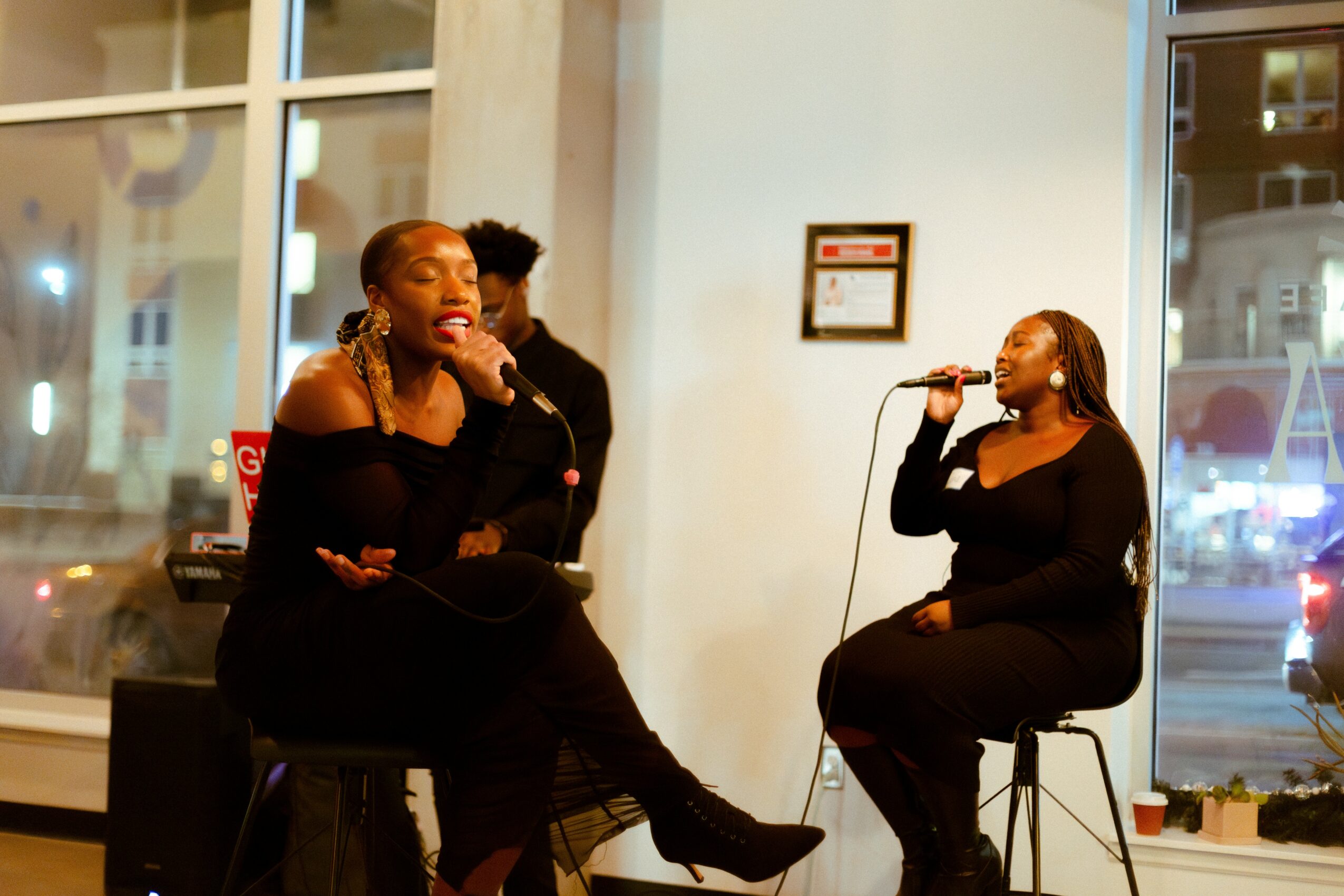 Two women performing on stage with a microphone, one sitting and singing, the other standing behind her, in a room with large windows.
