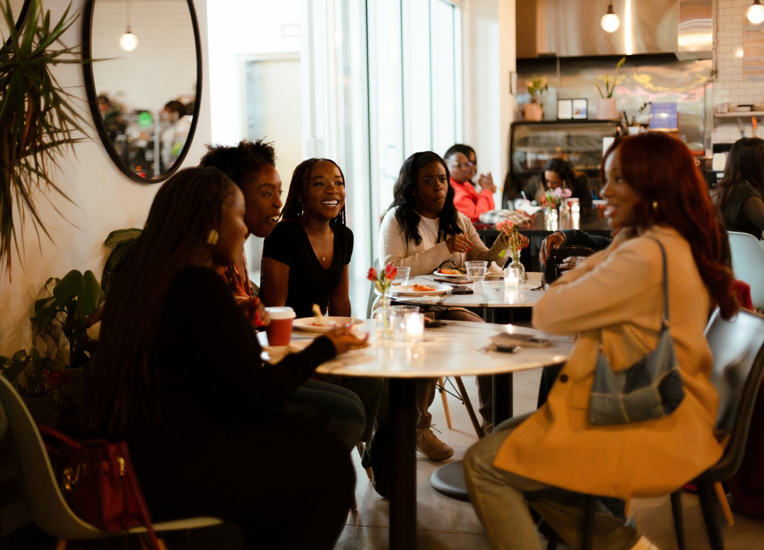Group of people sitting at a table in a cafe, engaging in conversation, with a window and kitchen in the background.