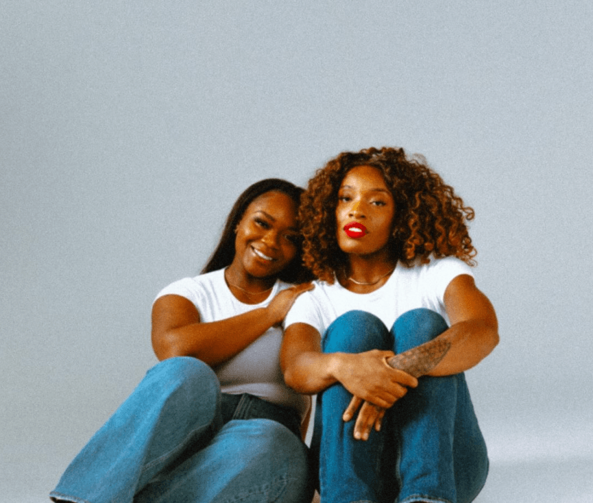 Two women sitting close together, smiling, against a plain light background.