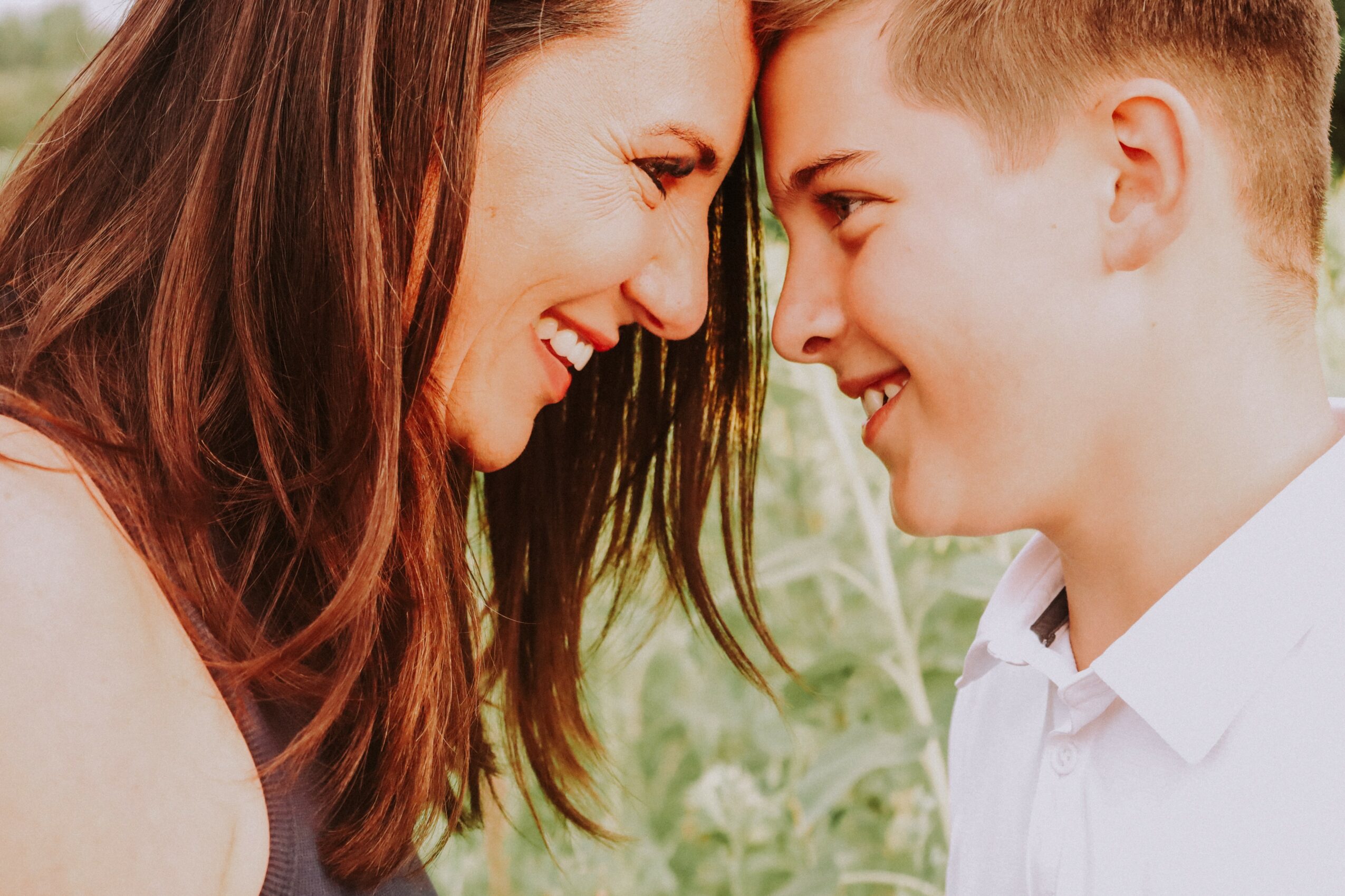 A woman and a young man face each other, smiling and touching foreheads outdoors.