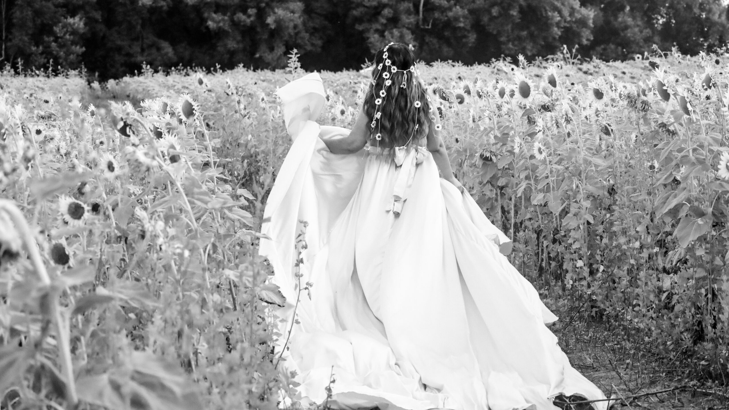 Woman in a flowing dress with flowers in her hair standing in a field of tall plants, holding a child.