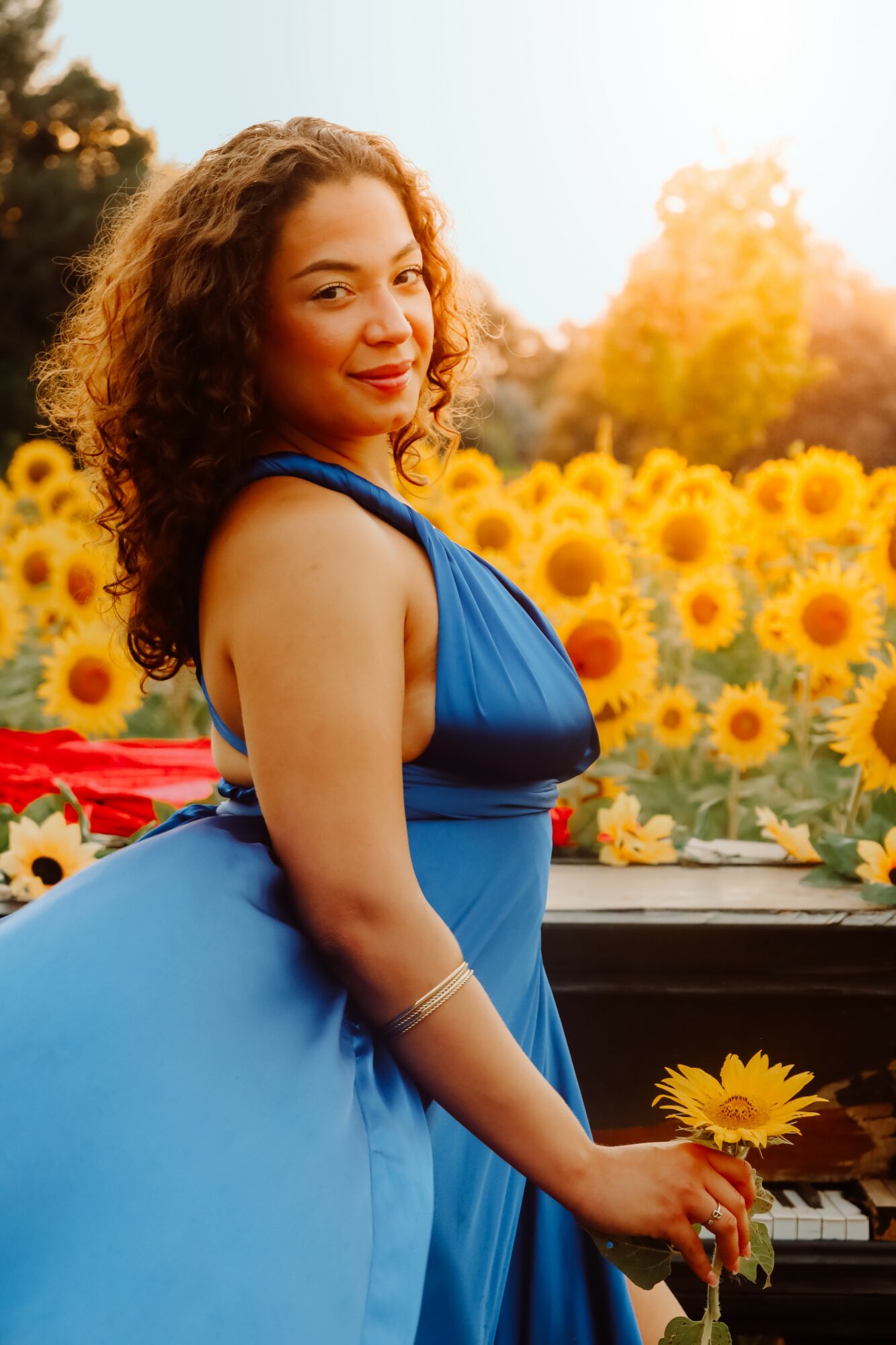 Young woman in blue dress standing in sunflower field during sunset, smiling, with trees in background.