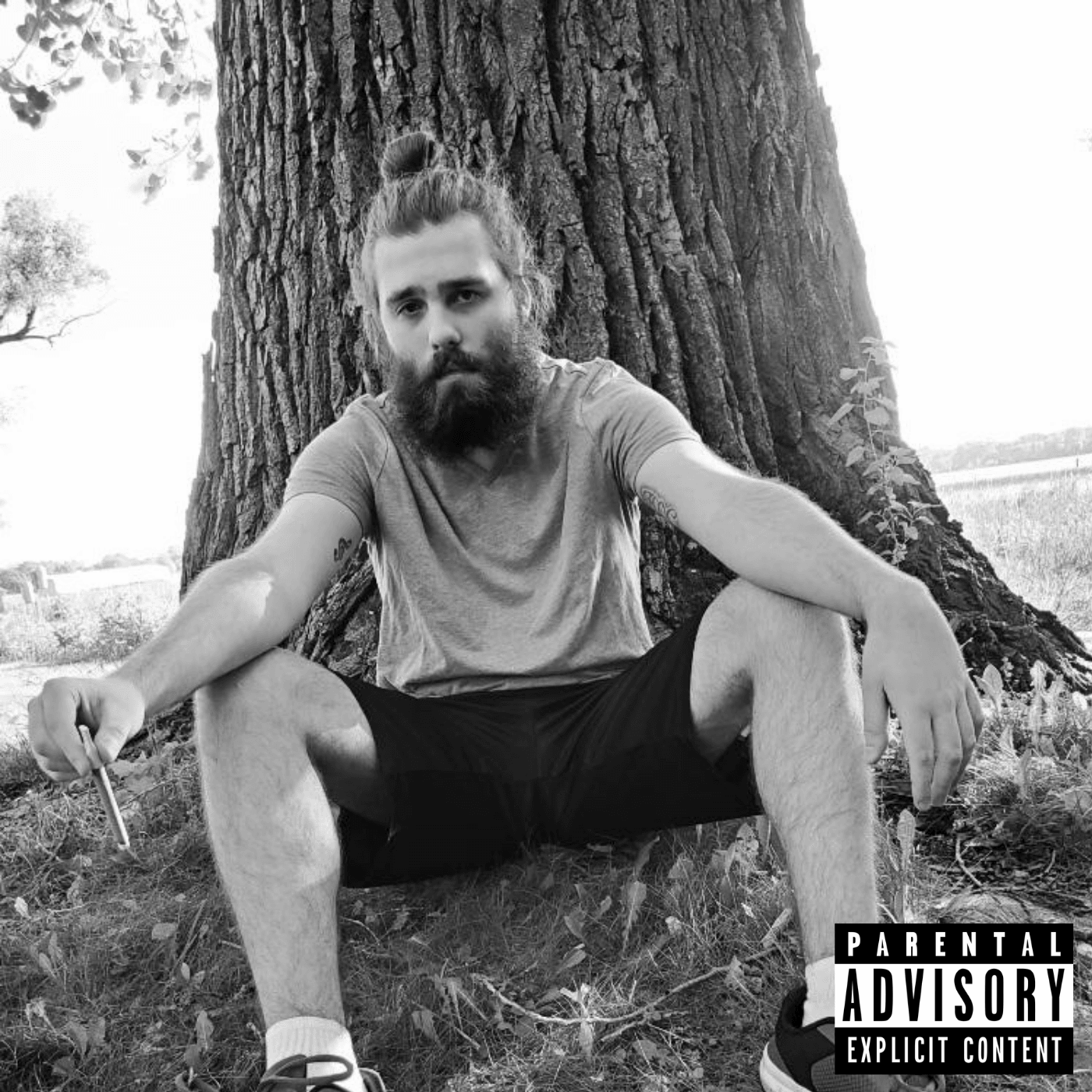 Person with a beard and bun sitting against a tree outdoors, wearing a t-shirt and shorts, holding a cigarette, in black and white.