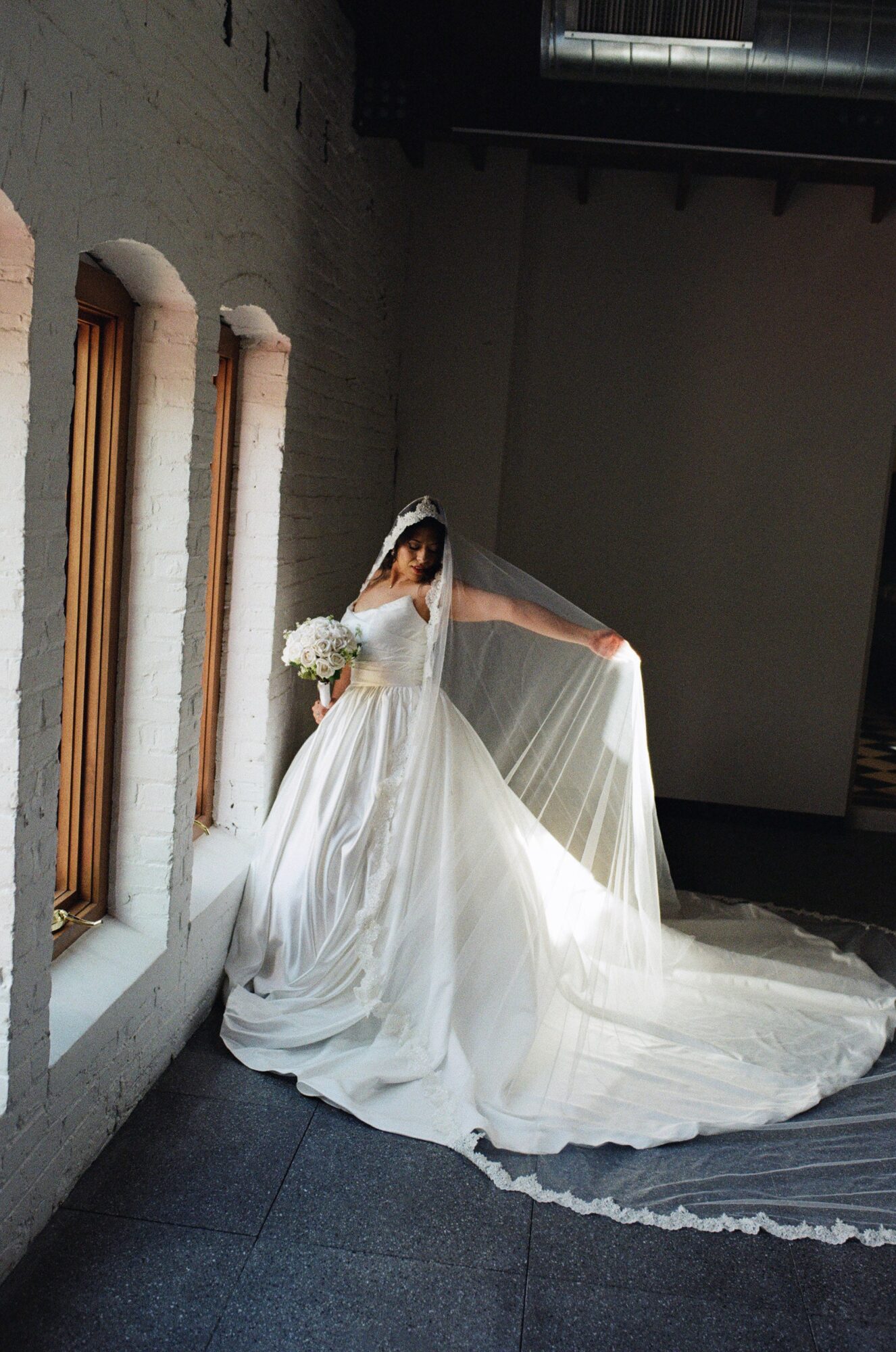 Bride in a white wedding dress with veil and bouquet standing by a window in a dimly lit room.