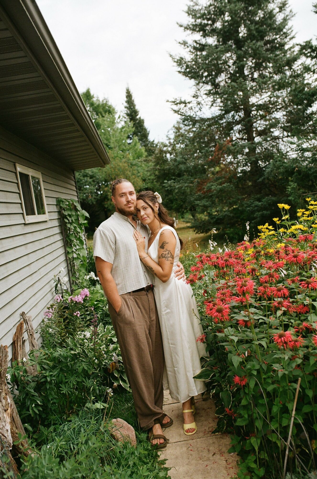 Two women stand in a garden with colorful flowers and trees, smiling and embracing each other.