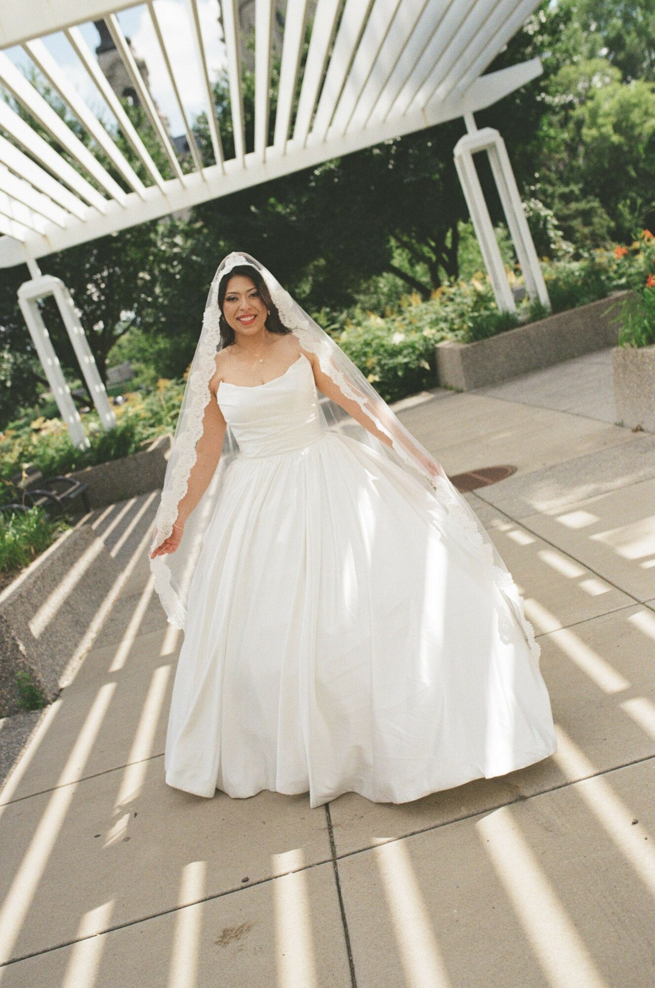 Woman in a white wedding dress standing outdoors on a wooden deck, with trees and a white structure in the background.