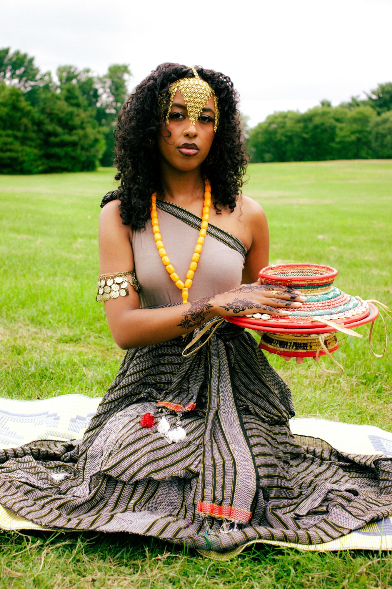 Woman sitting on grass holding colorful woven baskets, wearing traditional attire and jewelry, outdoors with trees and sky in background.