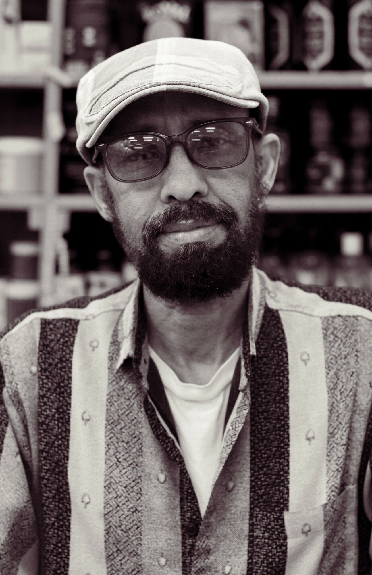 Man with glasses, beard, wearing a cap and patterned shirt, standing in front of shelves with bottles.