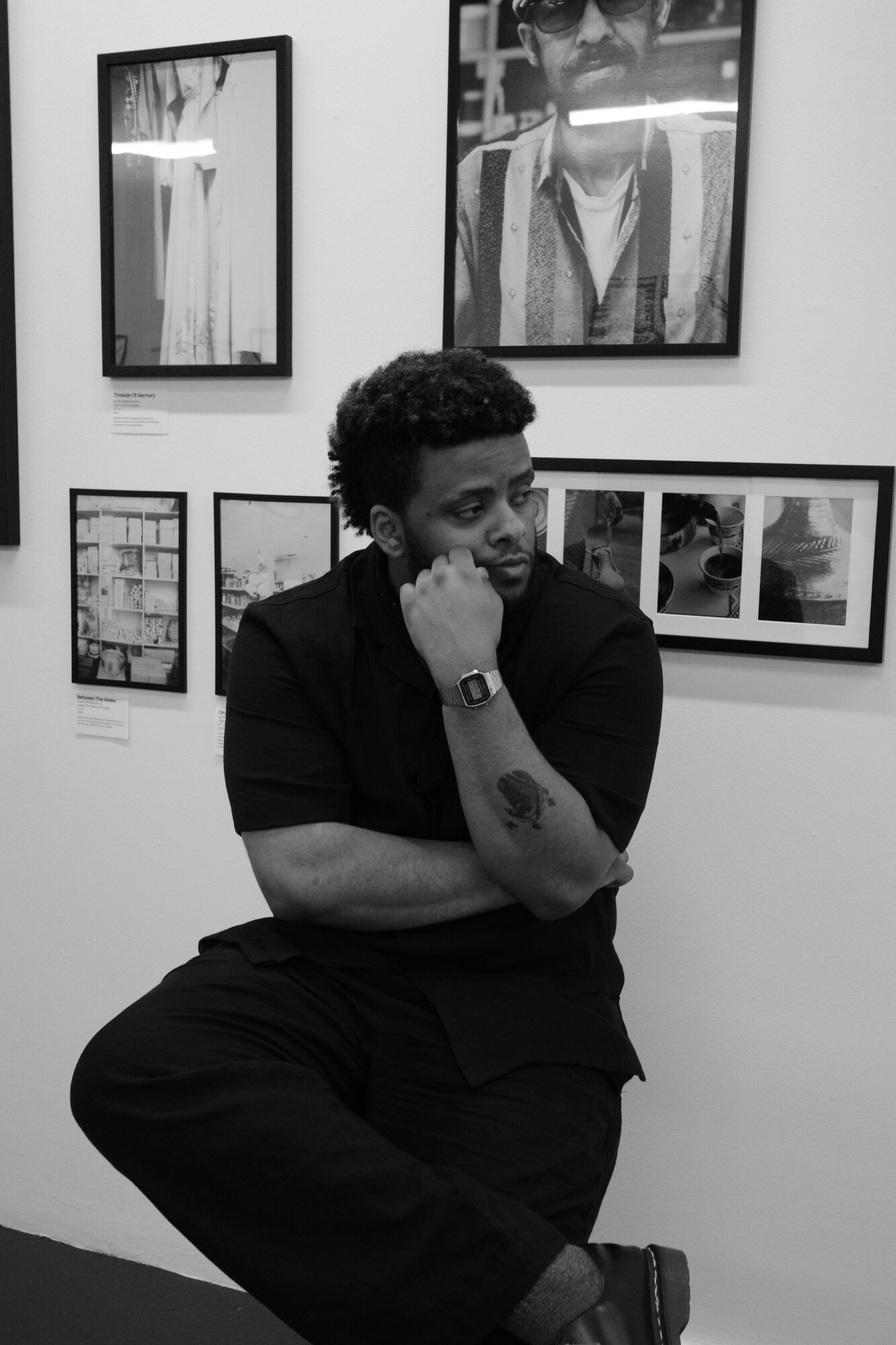 Person with curly hair and beard, wearing black, standing in front of framed pictures on a white wall, holding a phone to their ear.