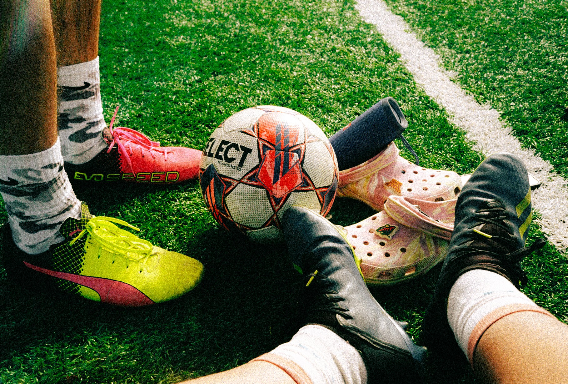 Soccer ball, shoes, and bags on a grassy field with white lines, two people sitting nearby, one with yellow shoes.