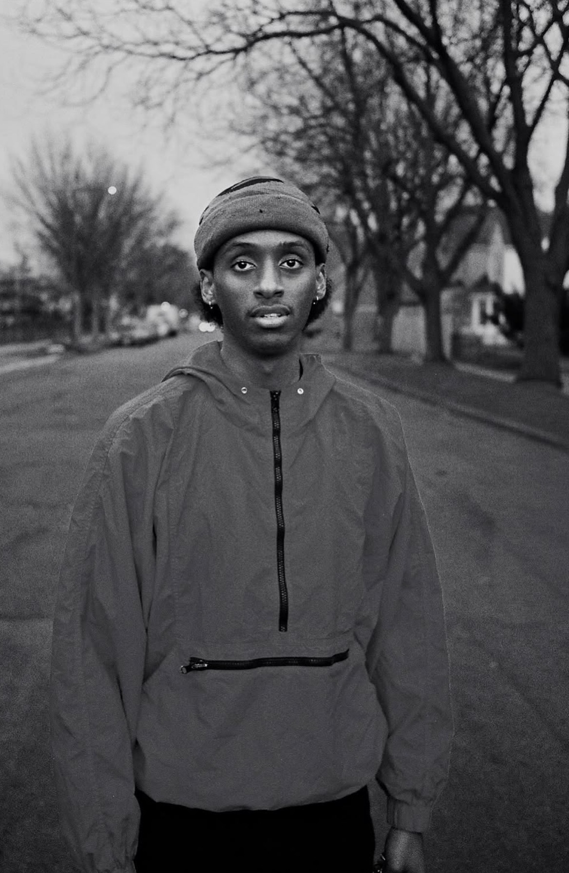 Young person wearing a headscarf and jacket standing outdoors on a tree-lined street, looking at the camera.