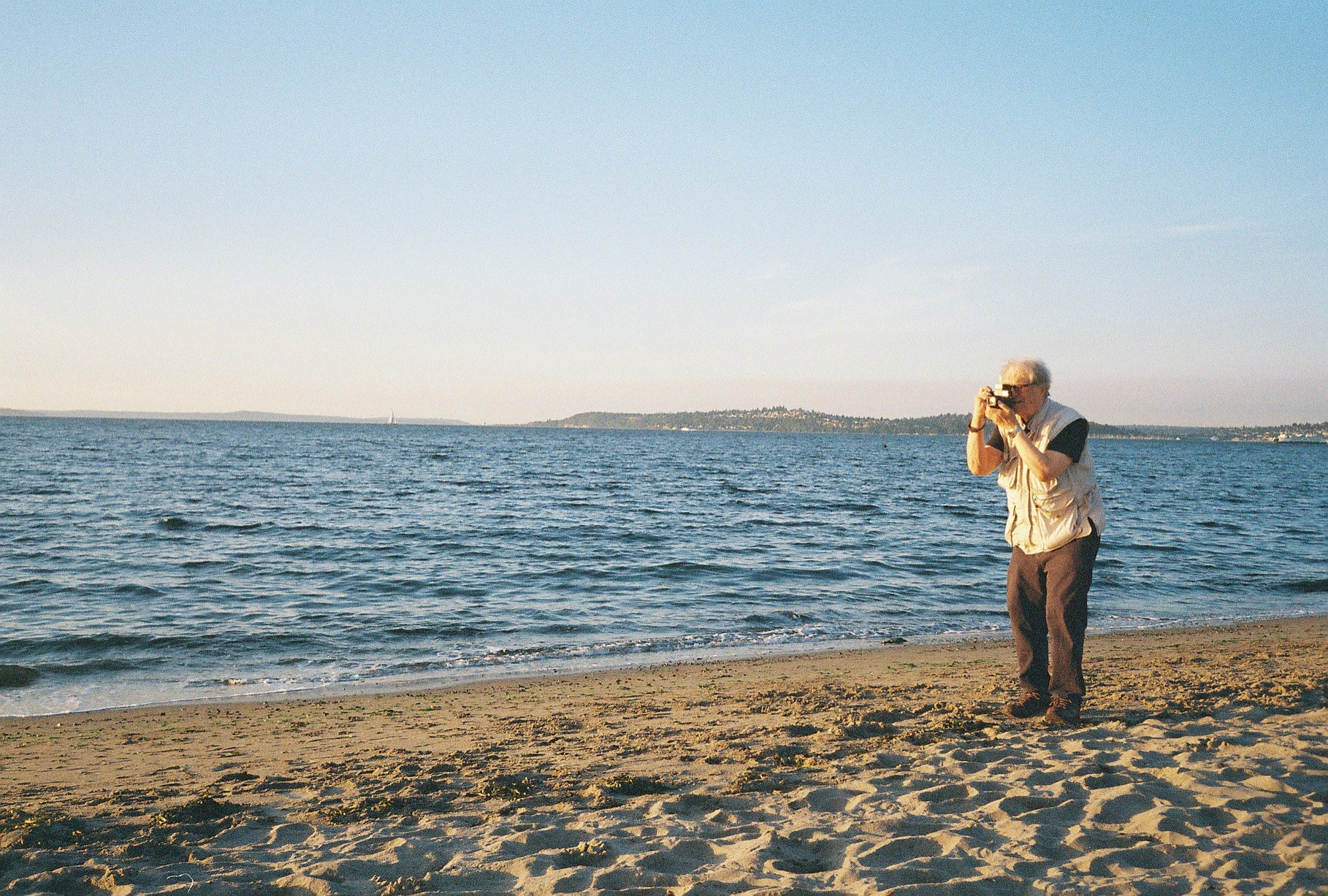 Person standing on a sandy beach taking a photo of the ocean with a camera, clear sky, distant land, and calm water