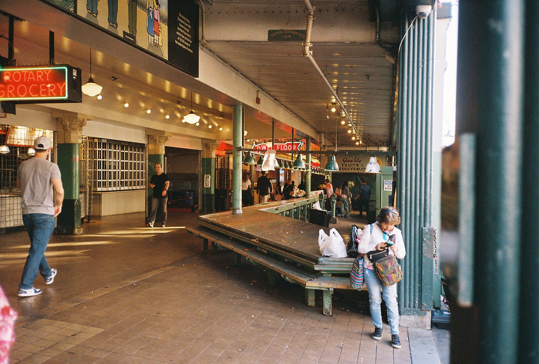 People walk and sit on benches inside a covered public space with shops and signs, some using phones or carrying bags.