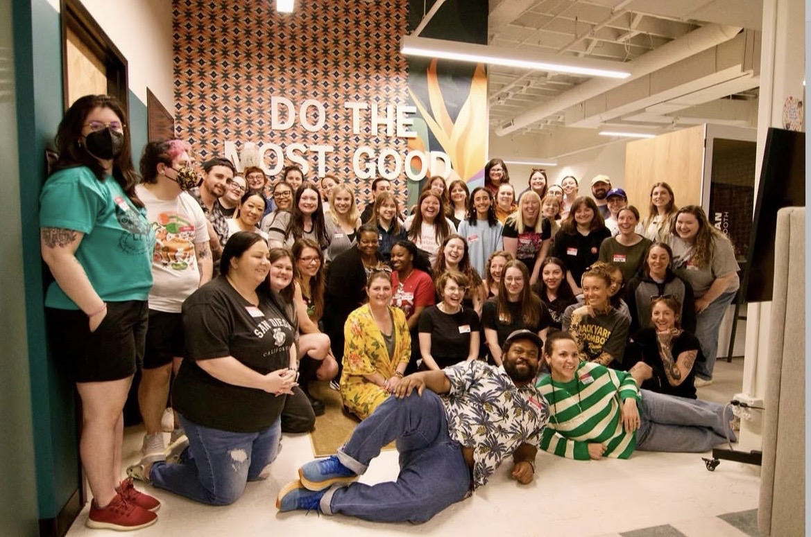 Group of people gathered indoors, some standing and some sitting, smiling for a photo, with a sign that says 'DO THE MOST GOOD'.