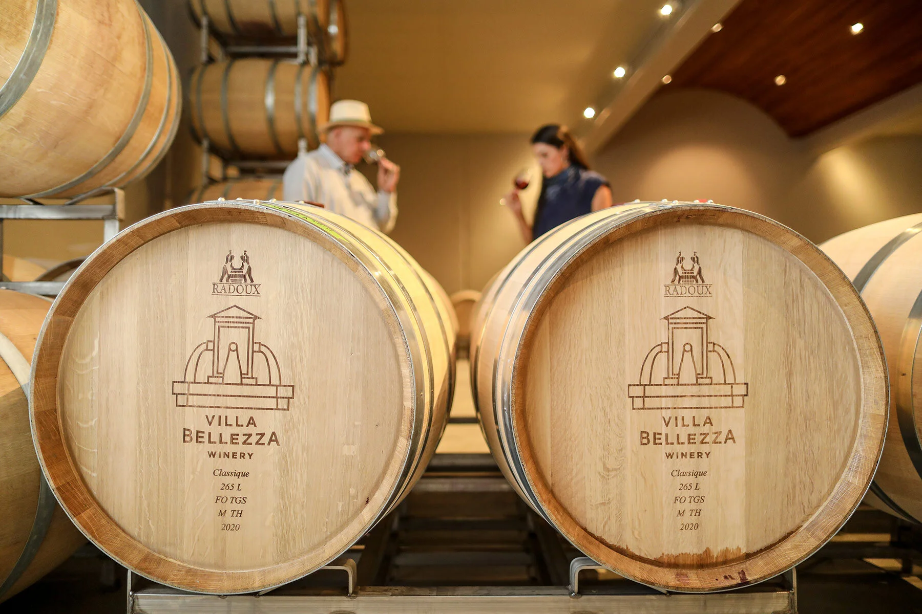 Two large wine barrels with Villa Bellezza logo in a winery, people in background, warm lighting.