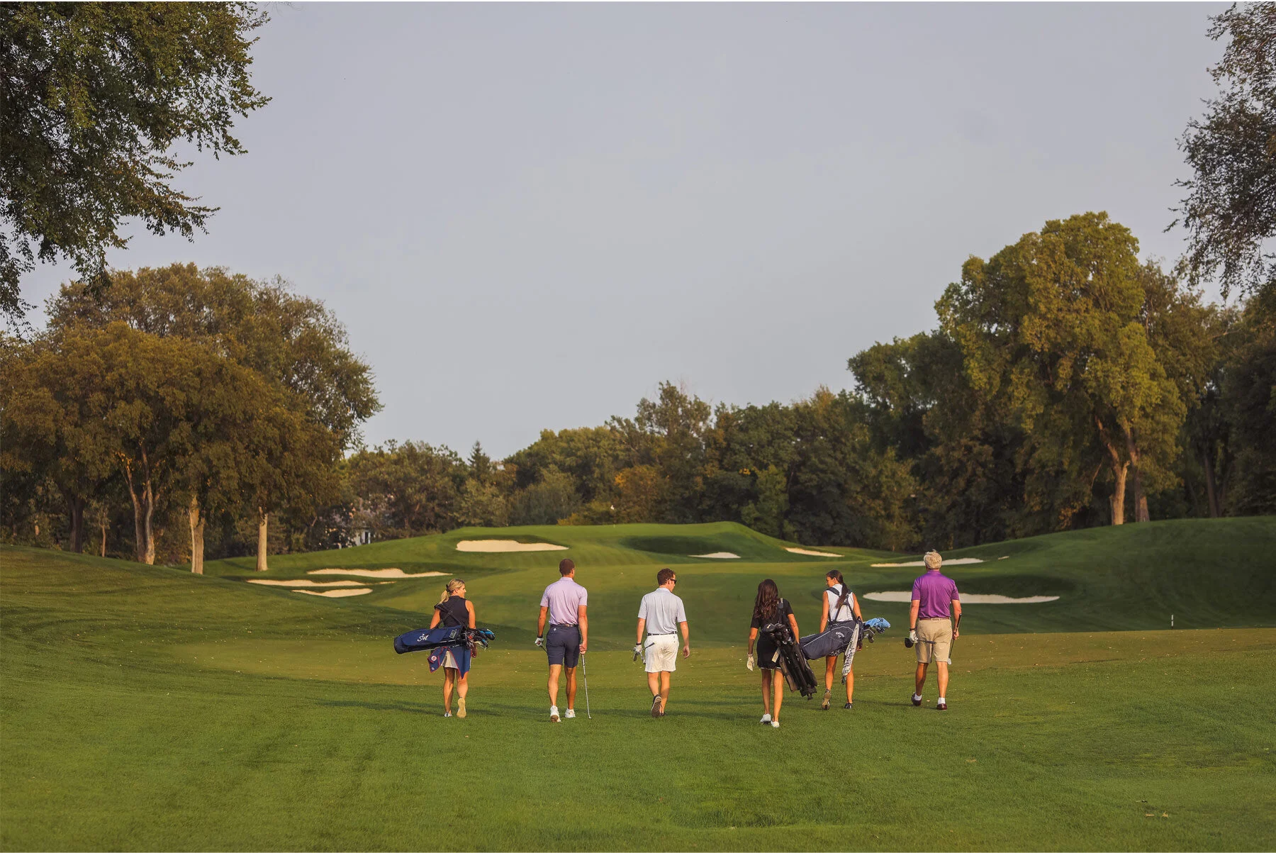 Group of seven people walking on a golf course fairway with trees and sand traps in the background.
