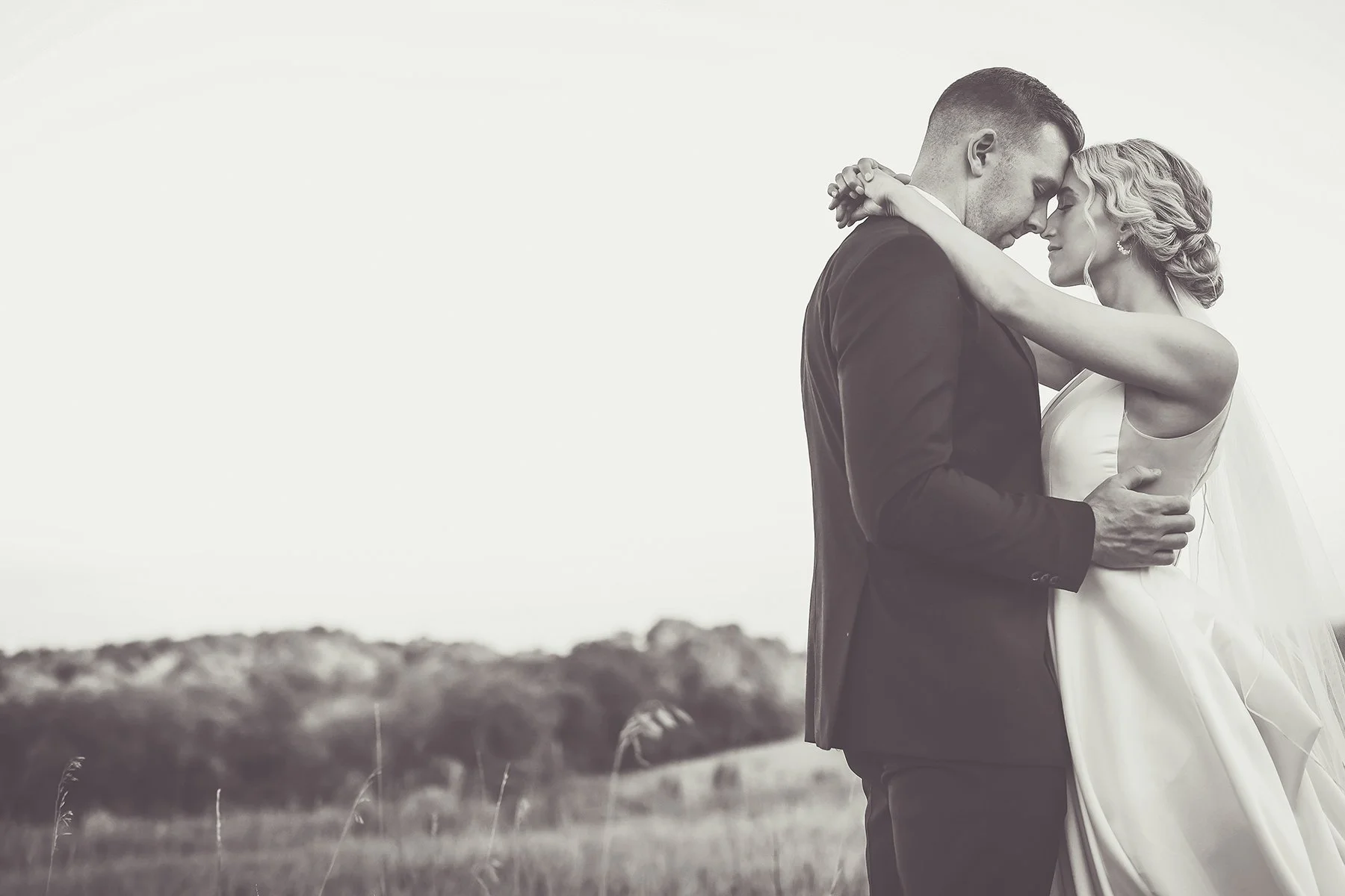 A couple in wedding attire embrace outdoors, facing each other with foreheads touching, in a natural landscape background.