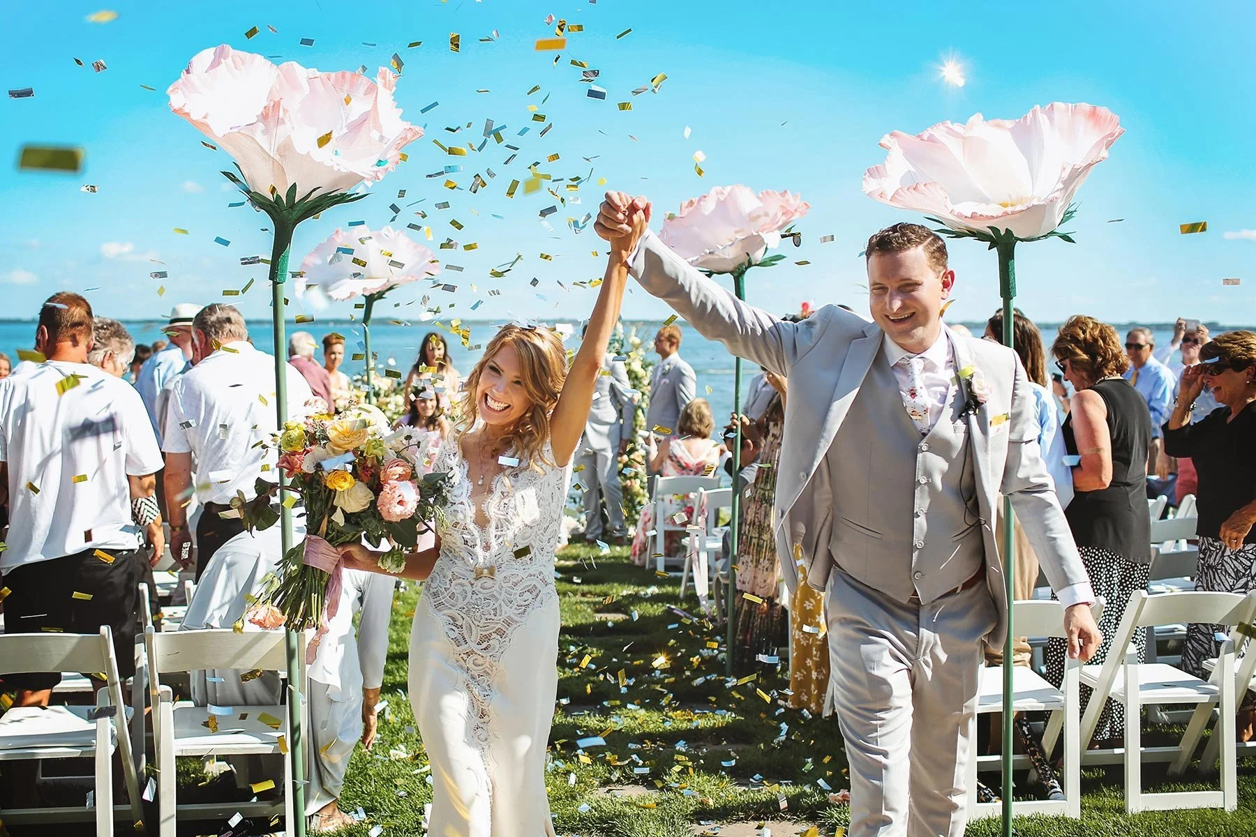 Bride and groom holding hands and celebrating outdoors with guests, flowers, and confetti, under a bright blue sky.