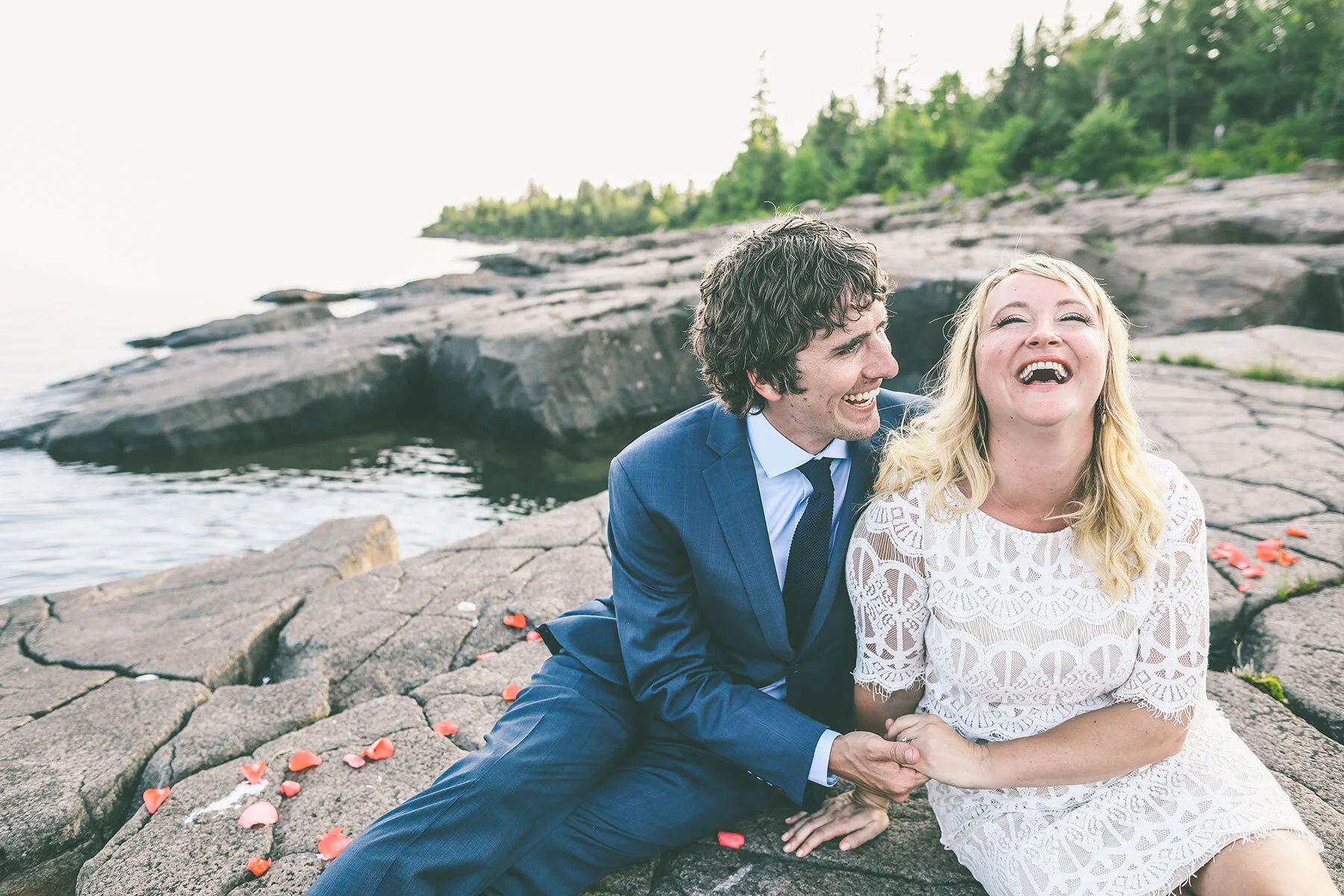Two people sitting on rocks near water, smiling and laughing, with trees in the background.
