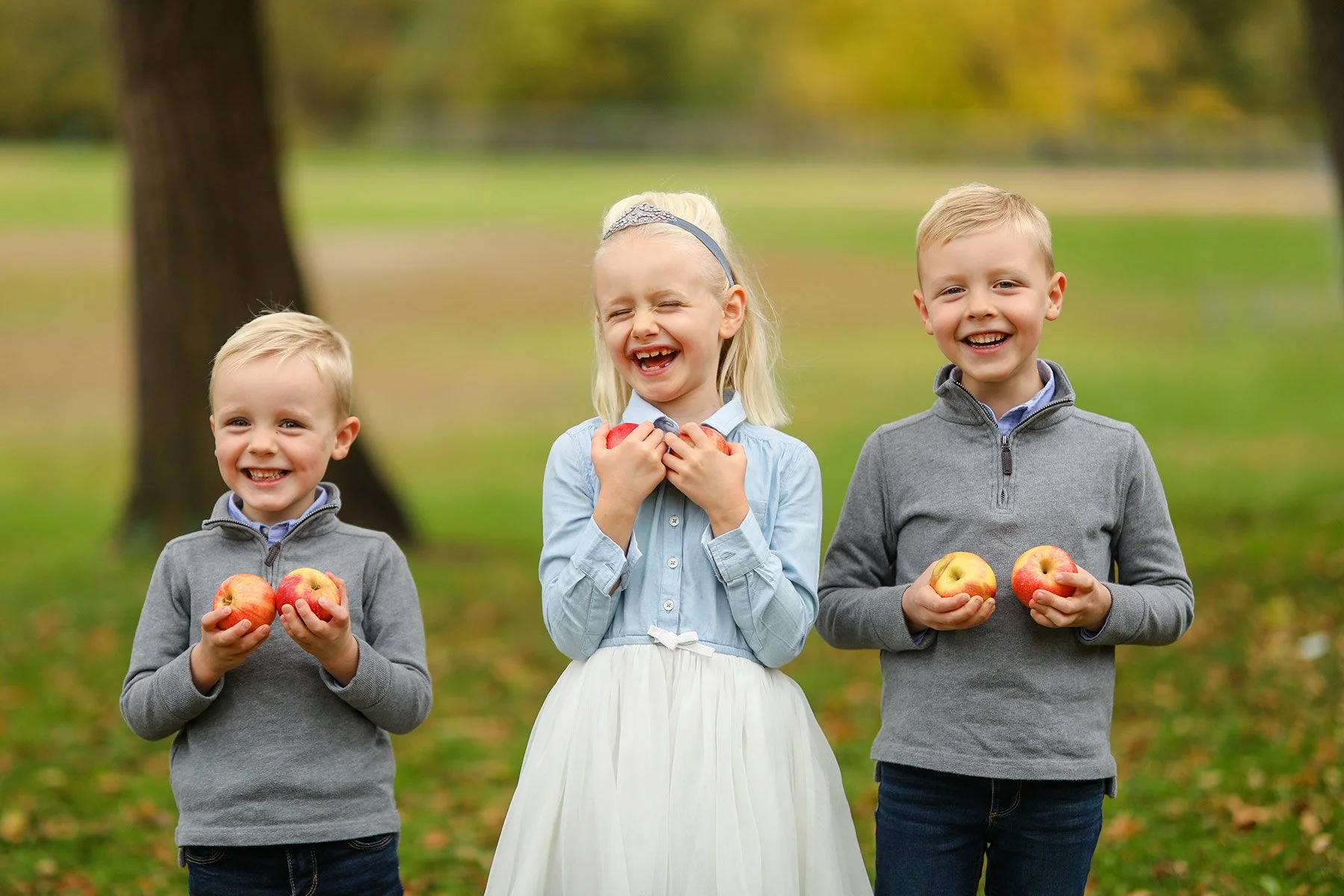 Three children smiling outdoors, holding apples, standing on grass with trees in background.