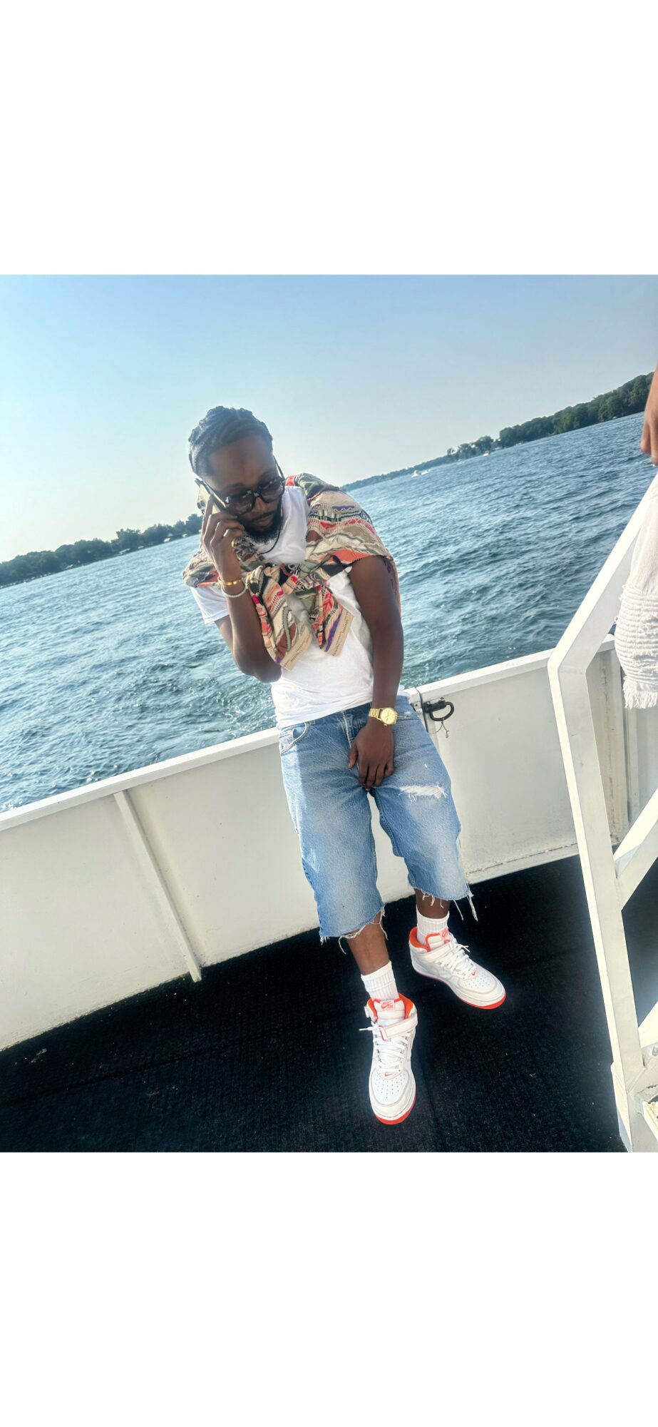 Person standing on boat with water and shoreline in background, wearing sunglasses, a patterned shirt, and jeans, holding a drink.