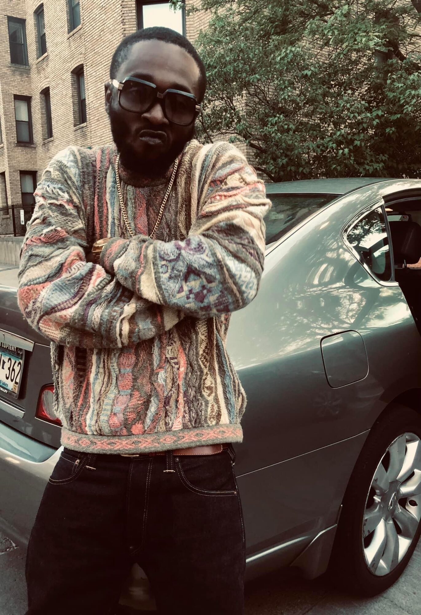 Man with glasses and beard wearing a colorful sweater, standing outdoors next to a car, arms crossed.